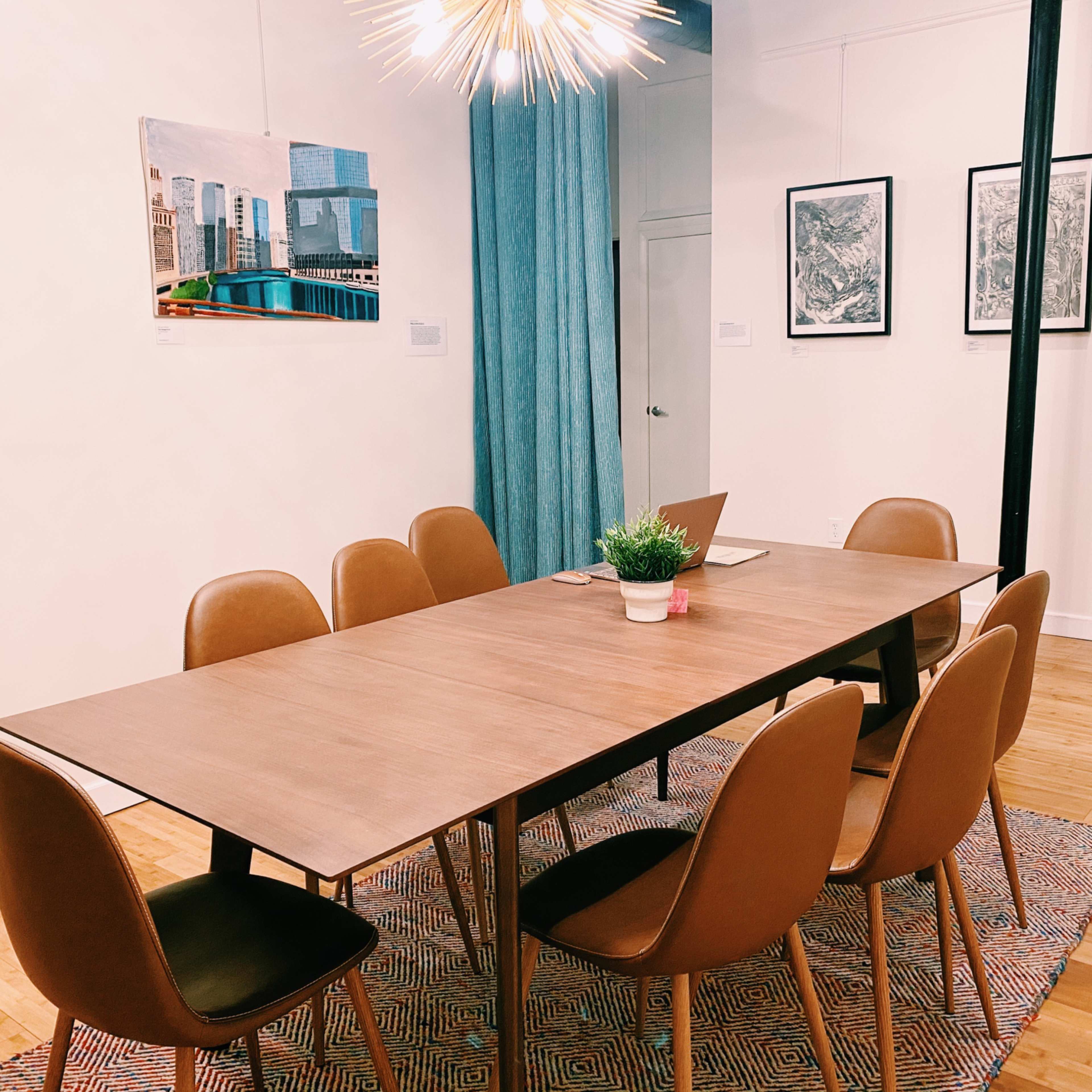 A wooden table surrounded by brown chairs is set in a well-lit room, with artwork displayed on the walls and a plant in the center.