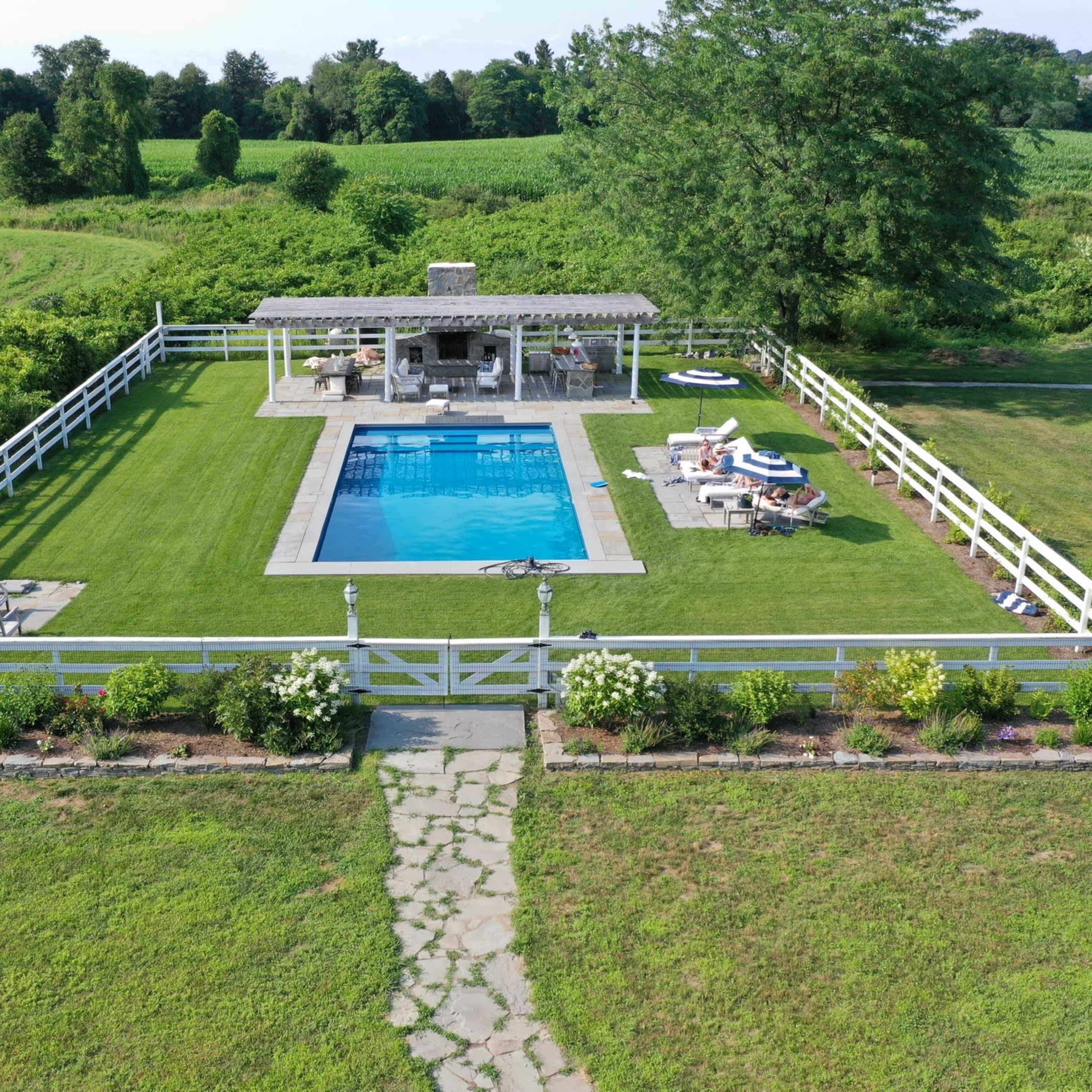 A fenced-in backyard with a swimming pool, a gazebo, and landscaped greenery surrounded by a large field.