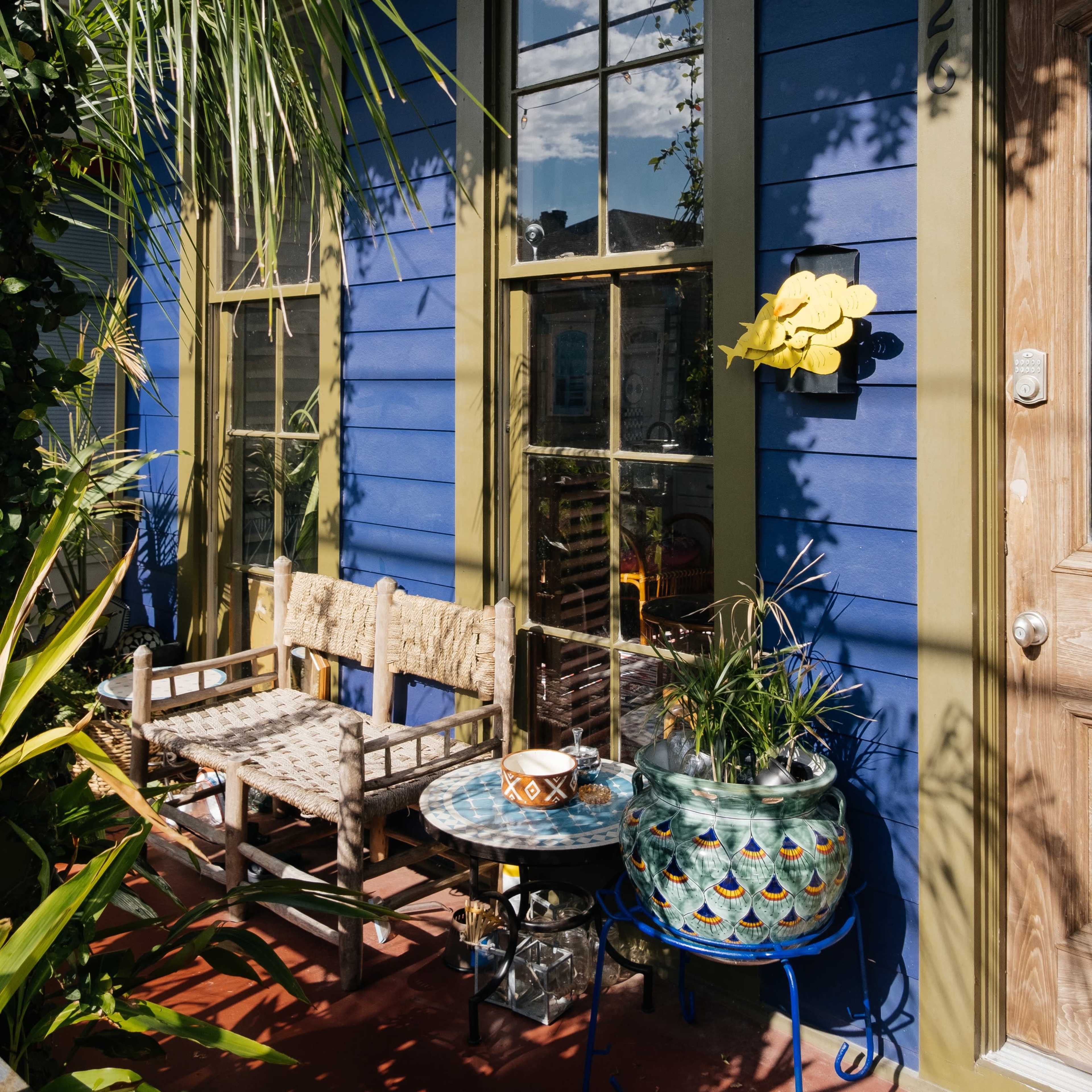 A vibrant blue house with large windows and a decorated porch, featuring a wooden bench, a table with colorful patterns, and various plants.