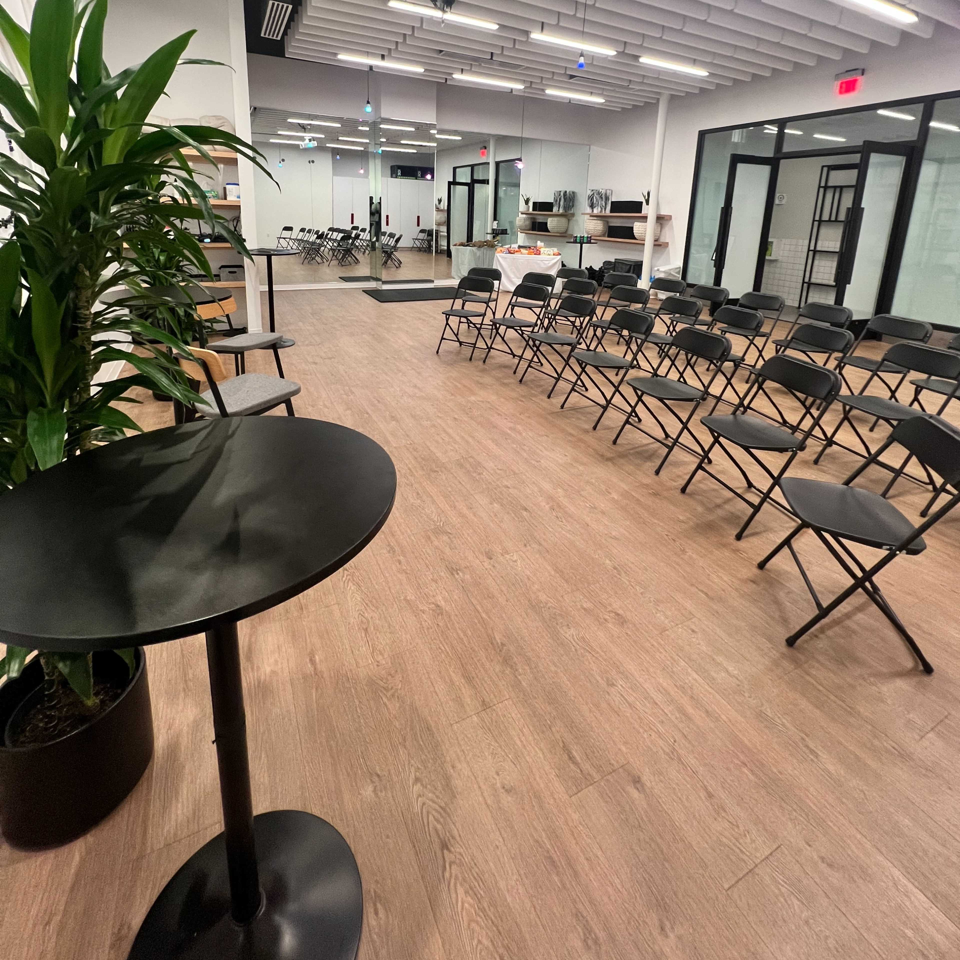 A spacious, modern meeting room with black folding chairs arranged in rows and a small round table with a plant beside it.
