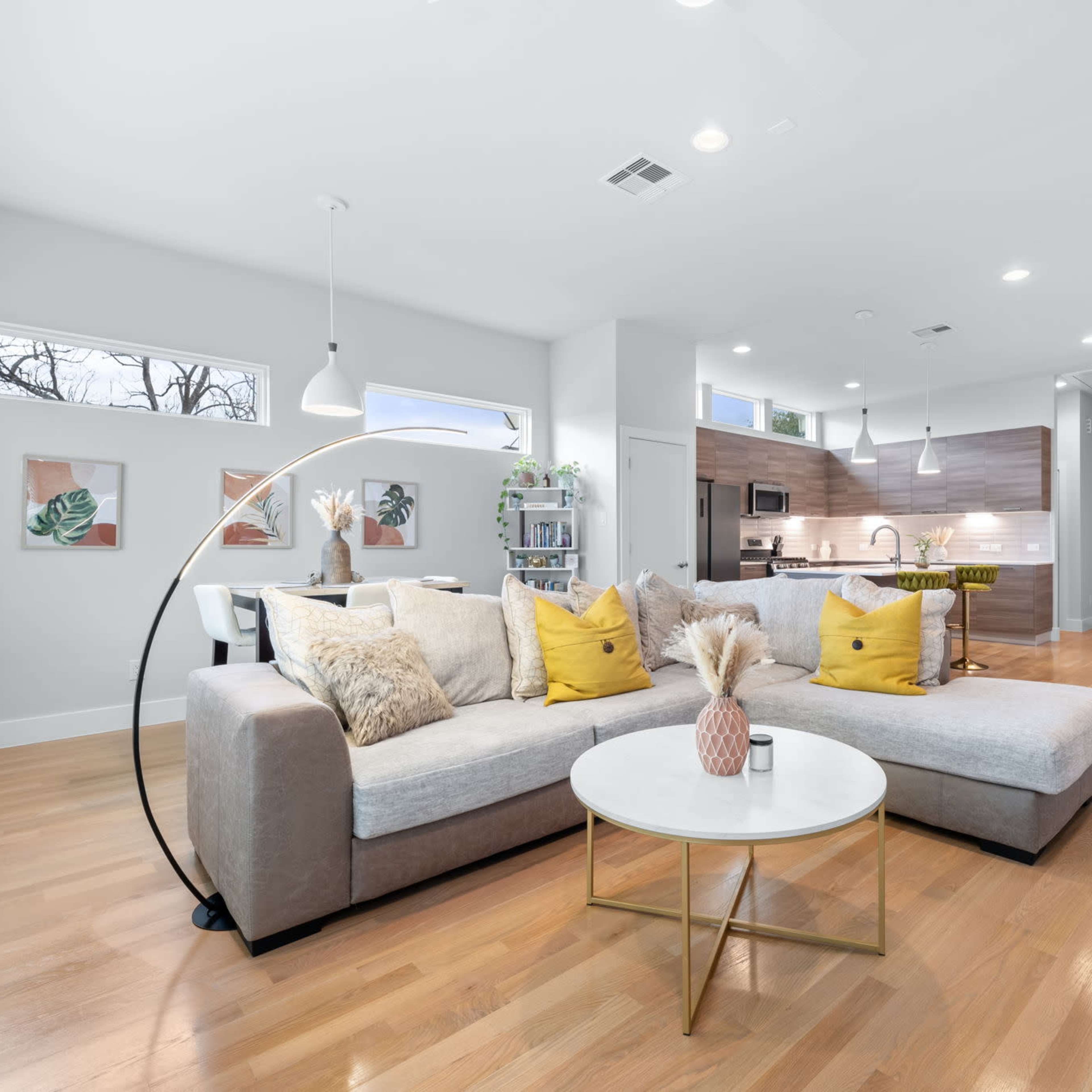 A modern living room features a gray sectional sofa with yellow accent pillows and a round coffee table, leading into a kitchen with wooden cabinetry.
