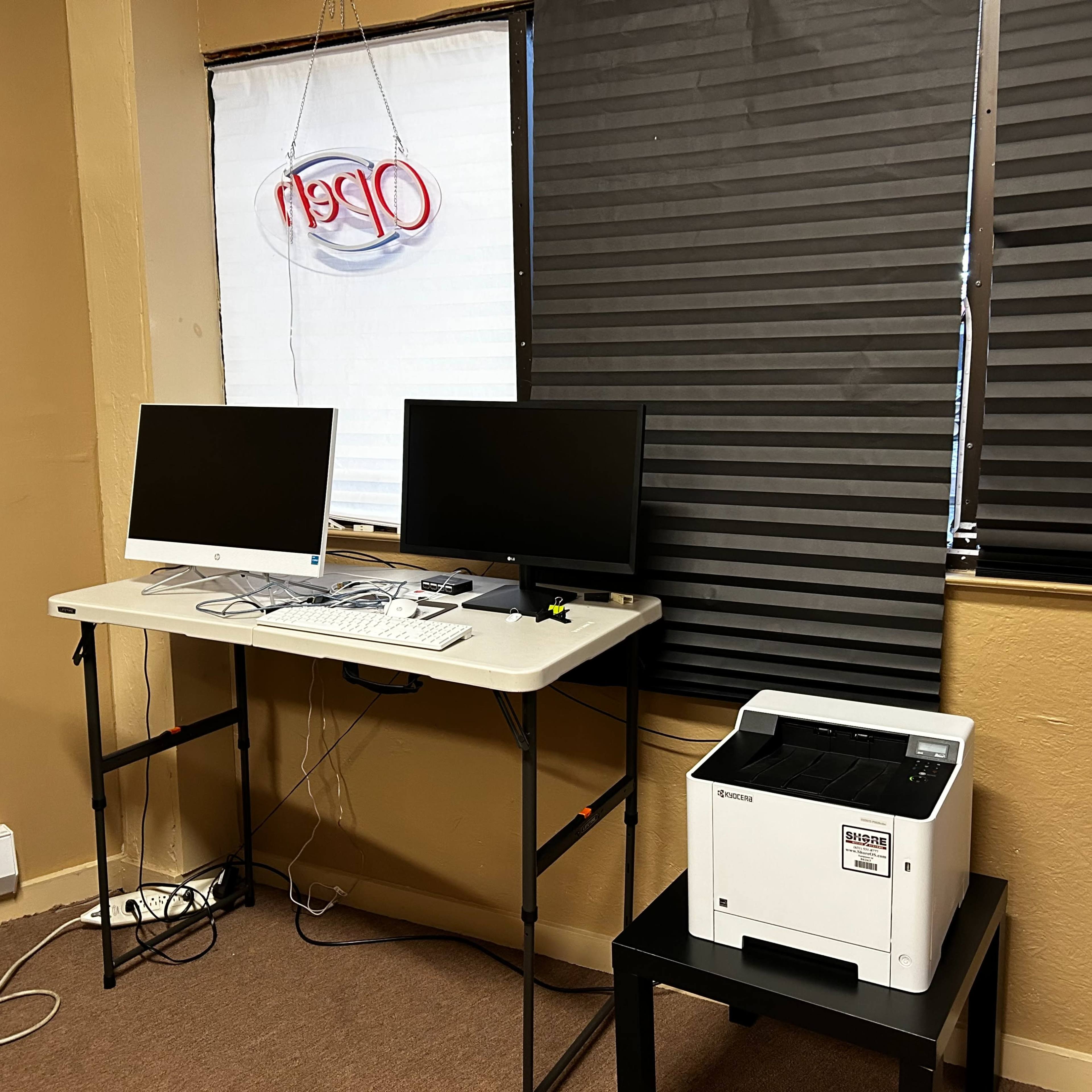 A small workspace features two computer monitors on a table, alongside a printer, with a window partially covered by blinds.