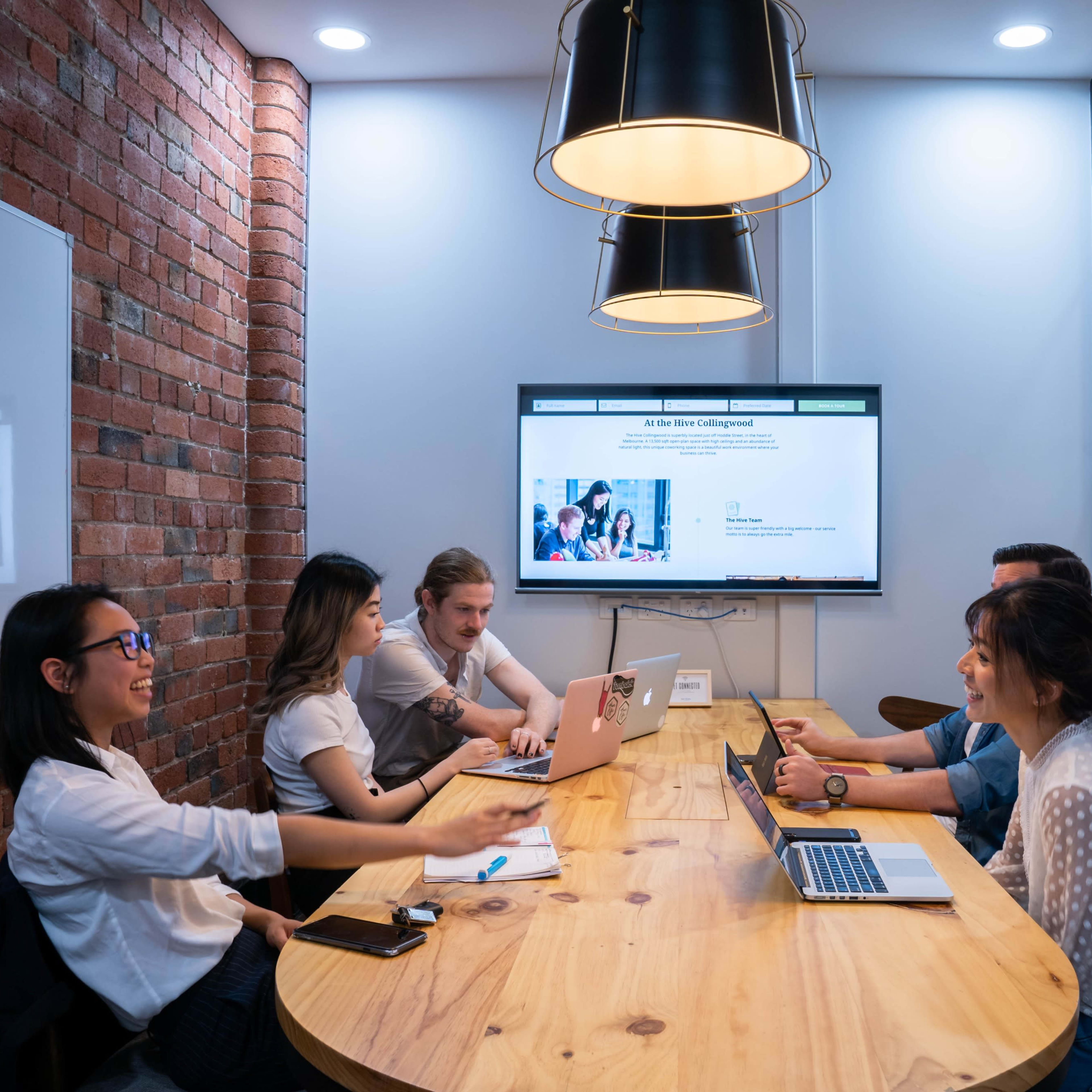 A group of five people are engaged in a meeting around a wooden table in a well-lit conference room with brick walls and a large screen displaying a presentation.