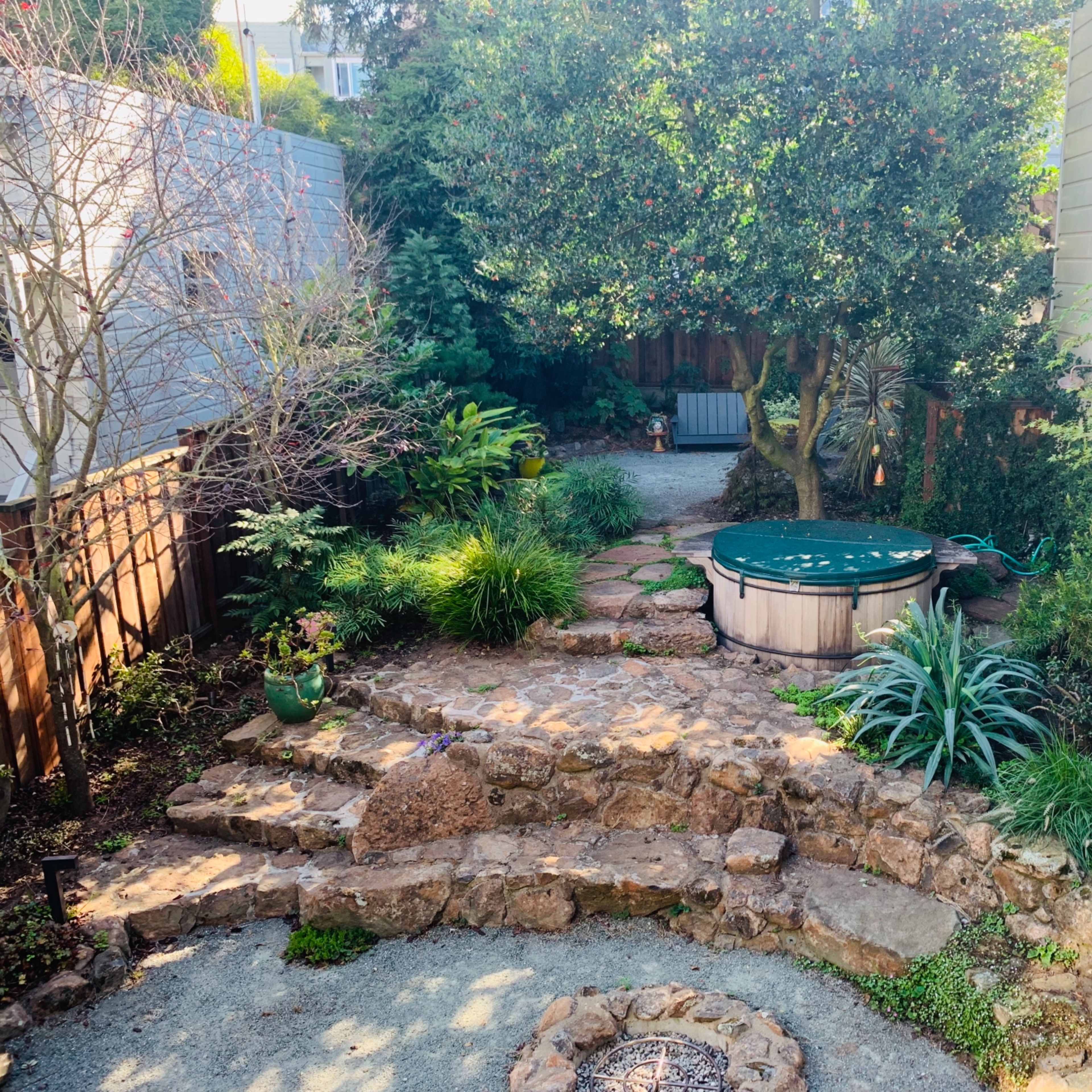 A landscaped garden featuring a circular hot tub surrounded by stone pathways, greenery, and a fire pit.