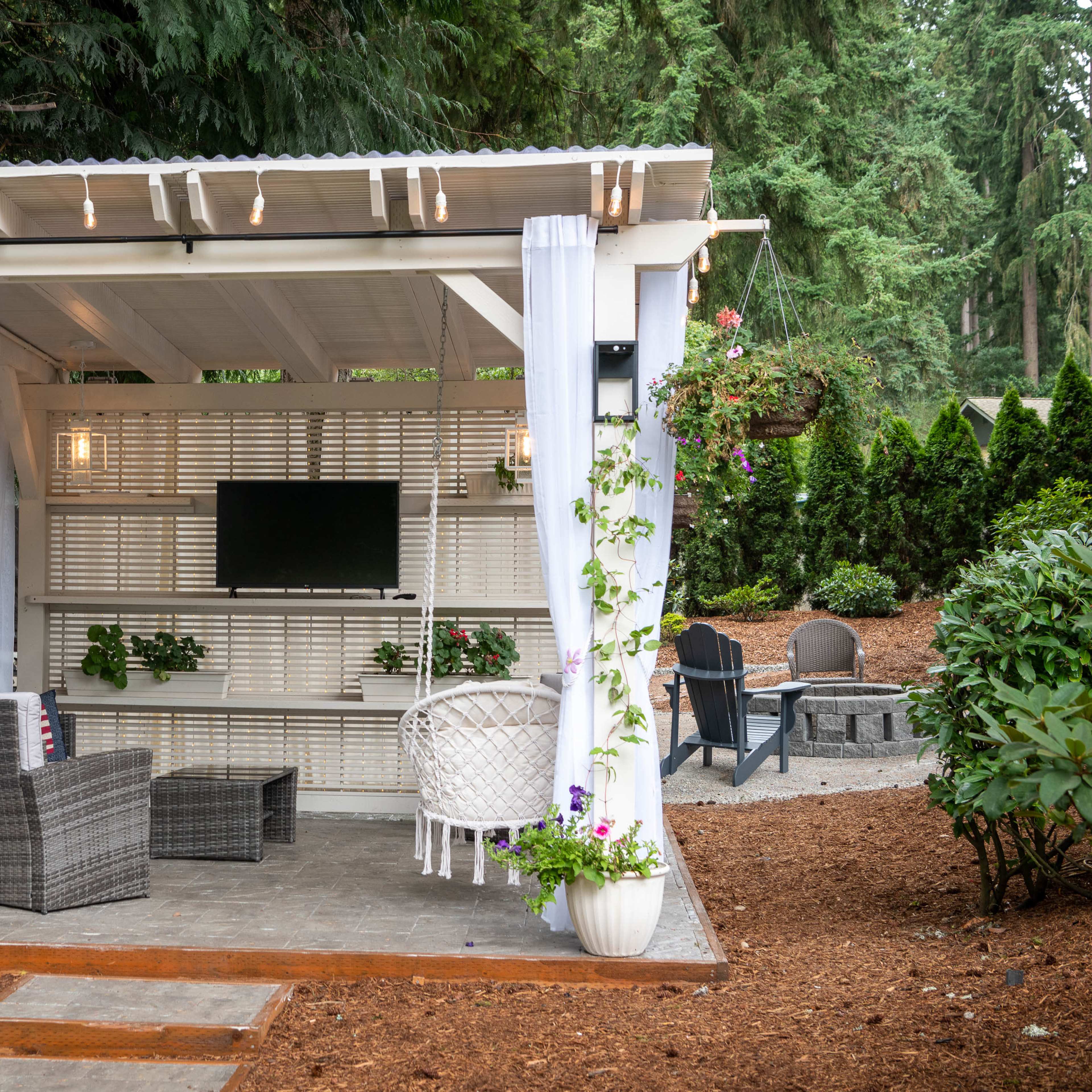 A cozy outdoor pavilion features comfortable seating, a wall-mounted television, and decorative plants, set against a backdrop of trees.