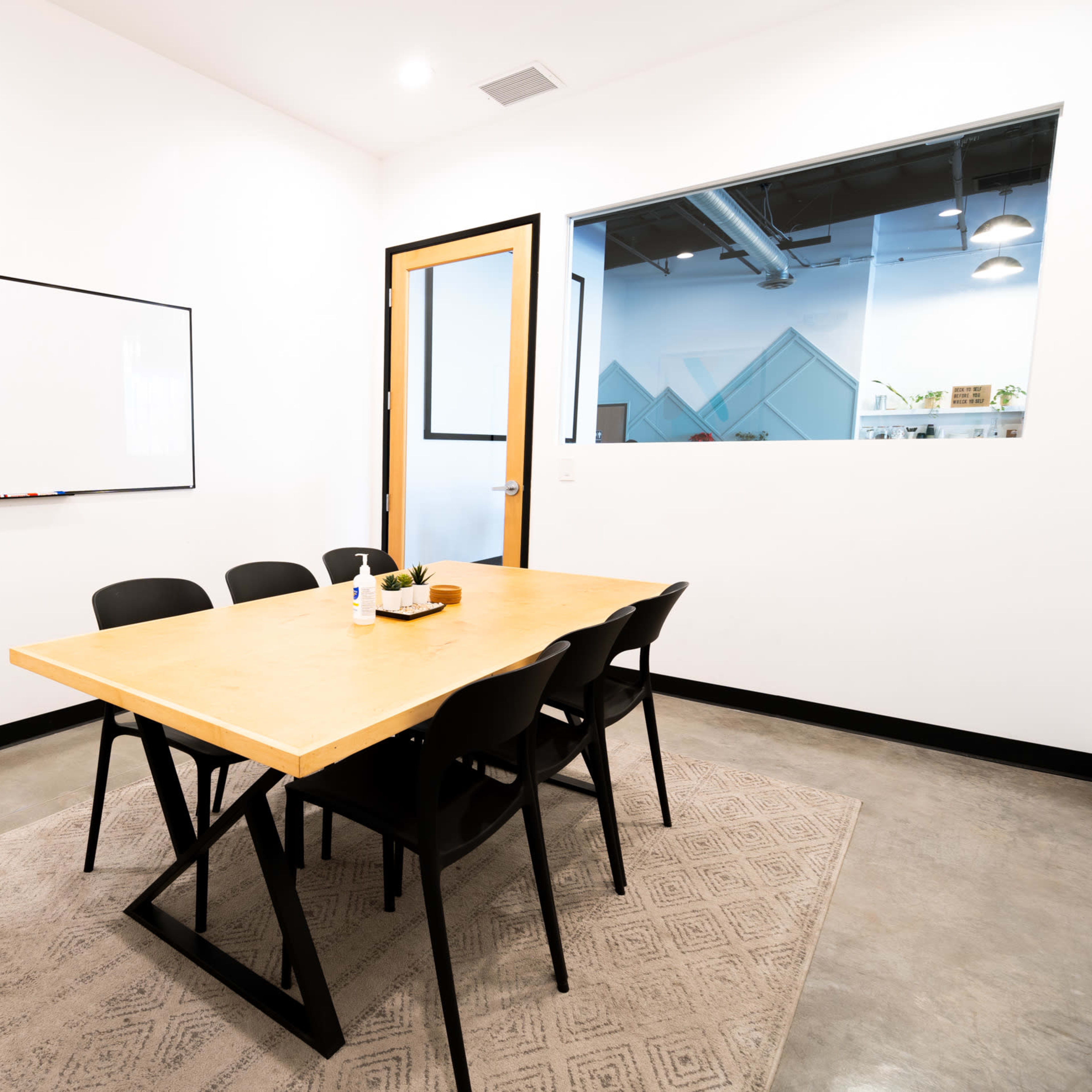 A meeting room with a rectangular wooden table and six black chairs, a whiteboard on one wall, and a glass door leading to another space.