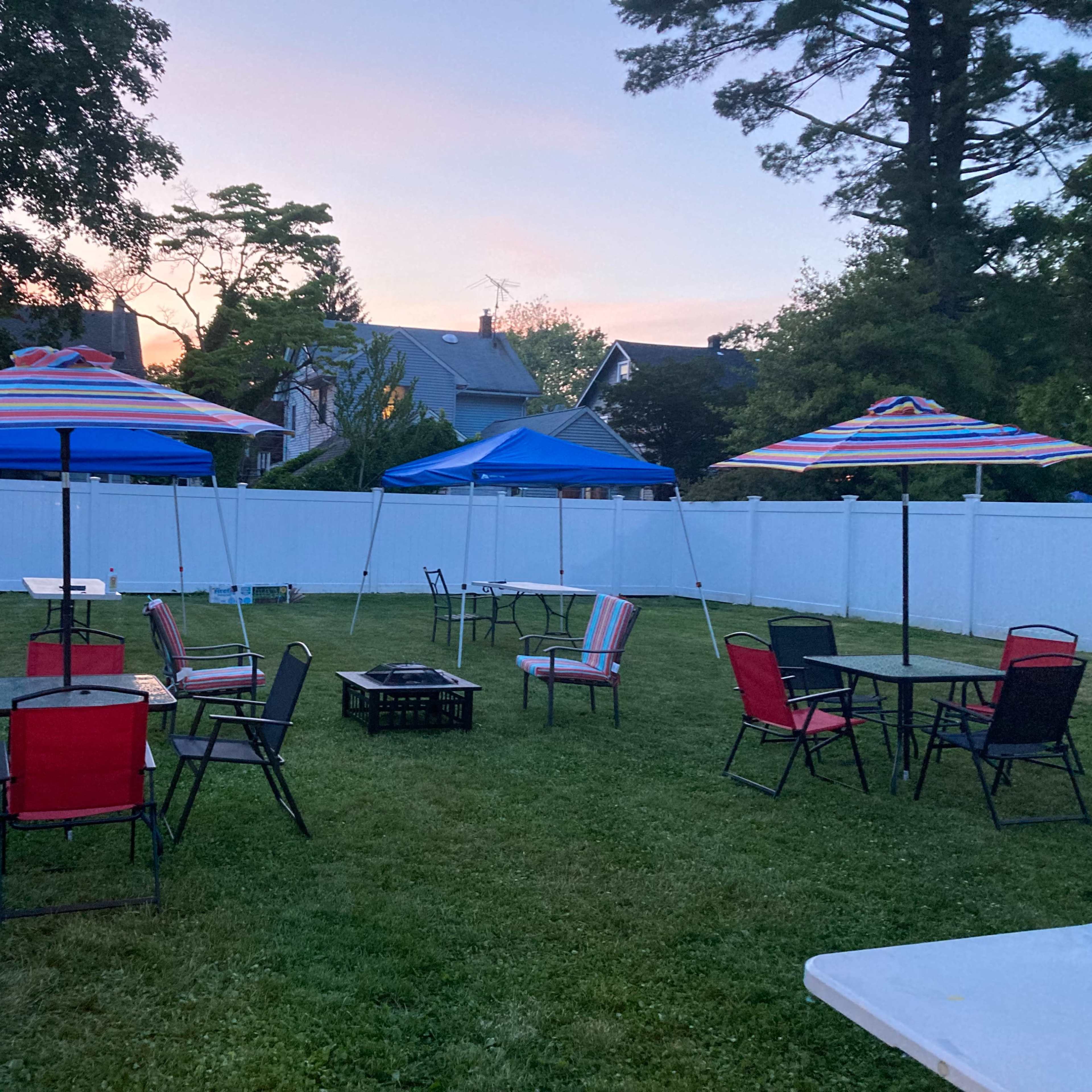 A grassy backyard with several tables and chairs under striped umbrellas, surrounded by a white fence at dusk.