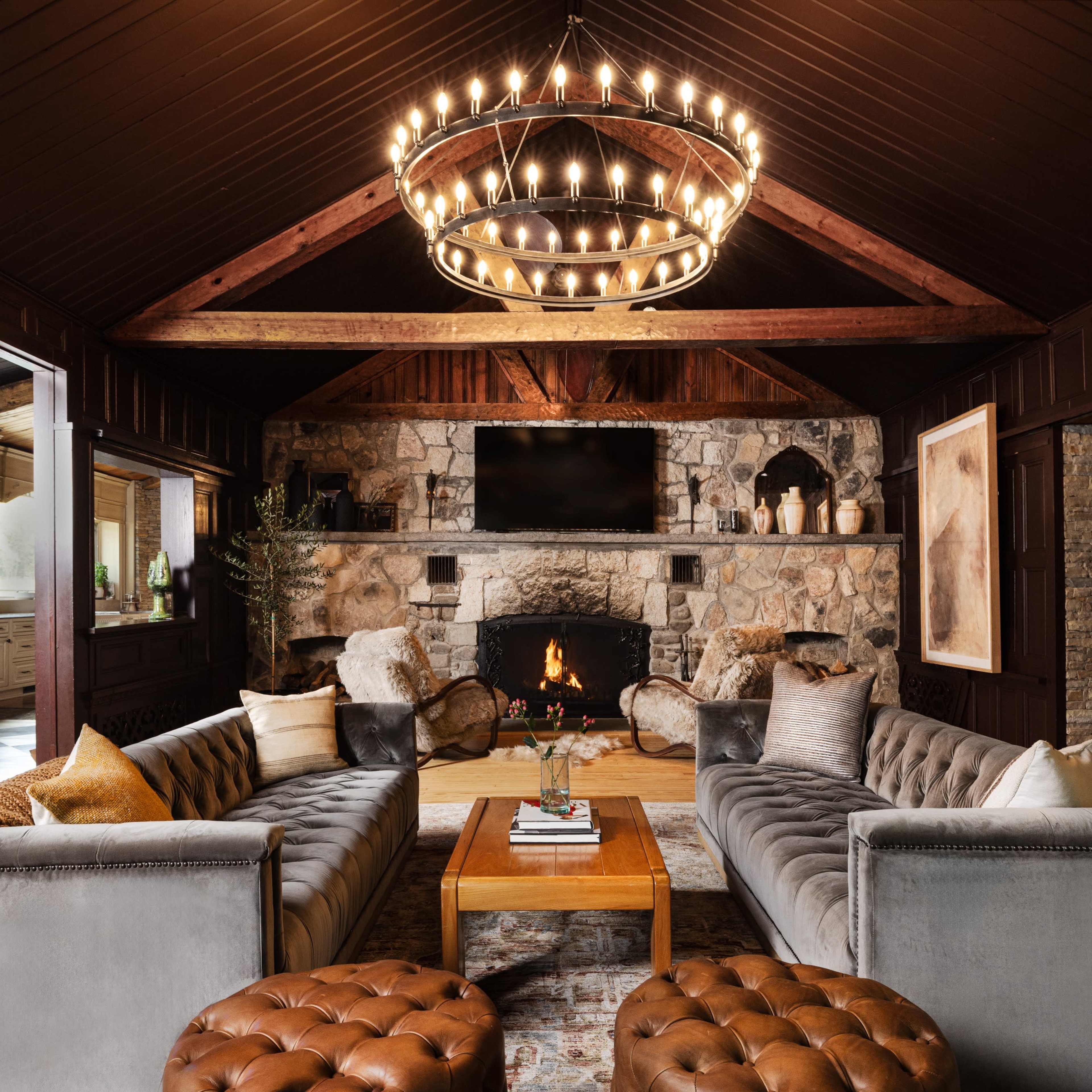 A cozy living room features two velvet sofas facing a stone fireplace, with a large chandelier overhead and wooden beams framing the ceiling.
