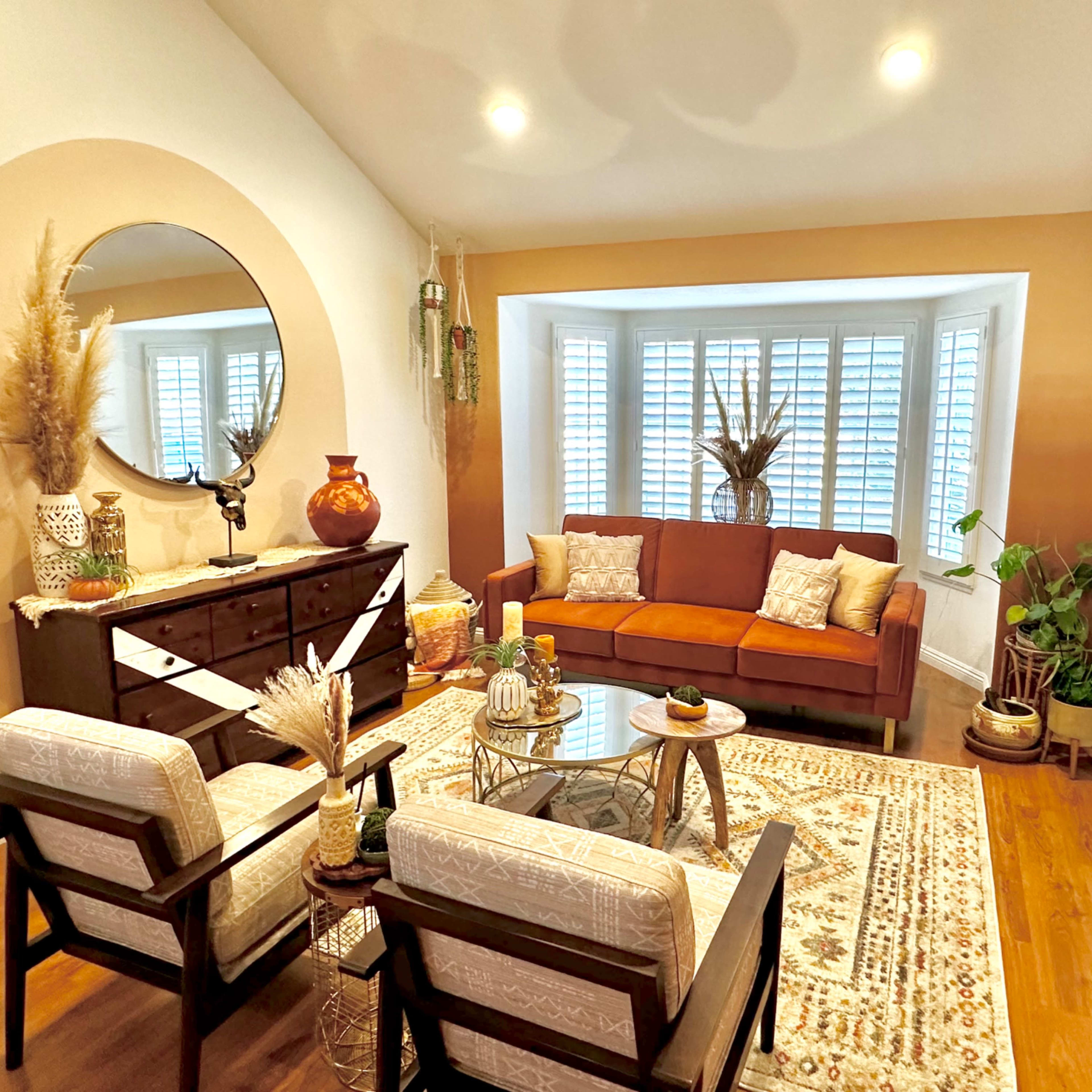 A cozy living room with a brown sofa, a glass coffee table, and decorative plants against a backdrop of white window shutters and accent wall decor.