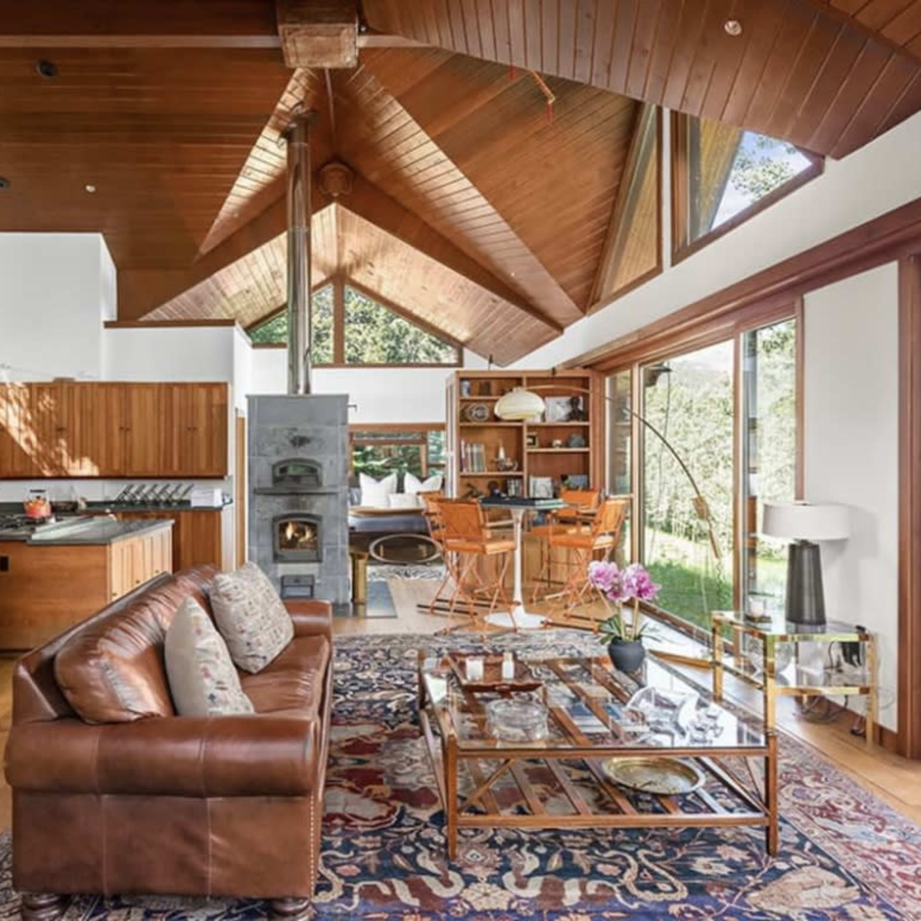 A spacious, modern living room with wooden ceilings, large windows, a brown leather sofa, and a dining area in the background.