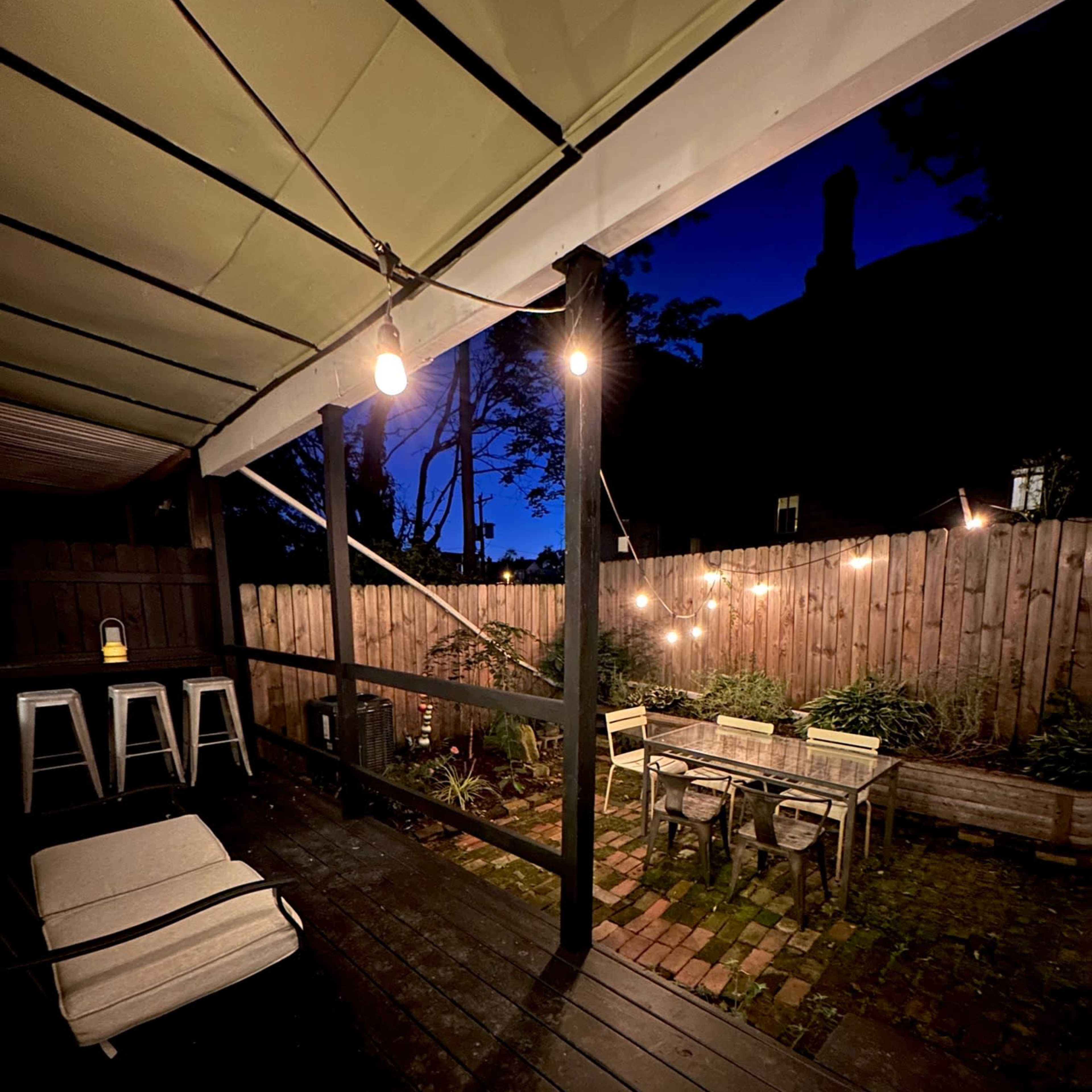 A dimly lit backyard patio with wooden furniture and string lights against a twilight sky.