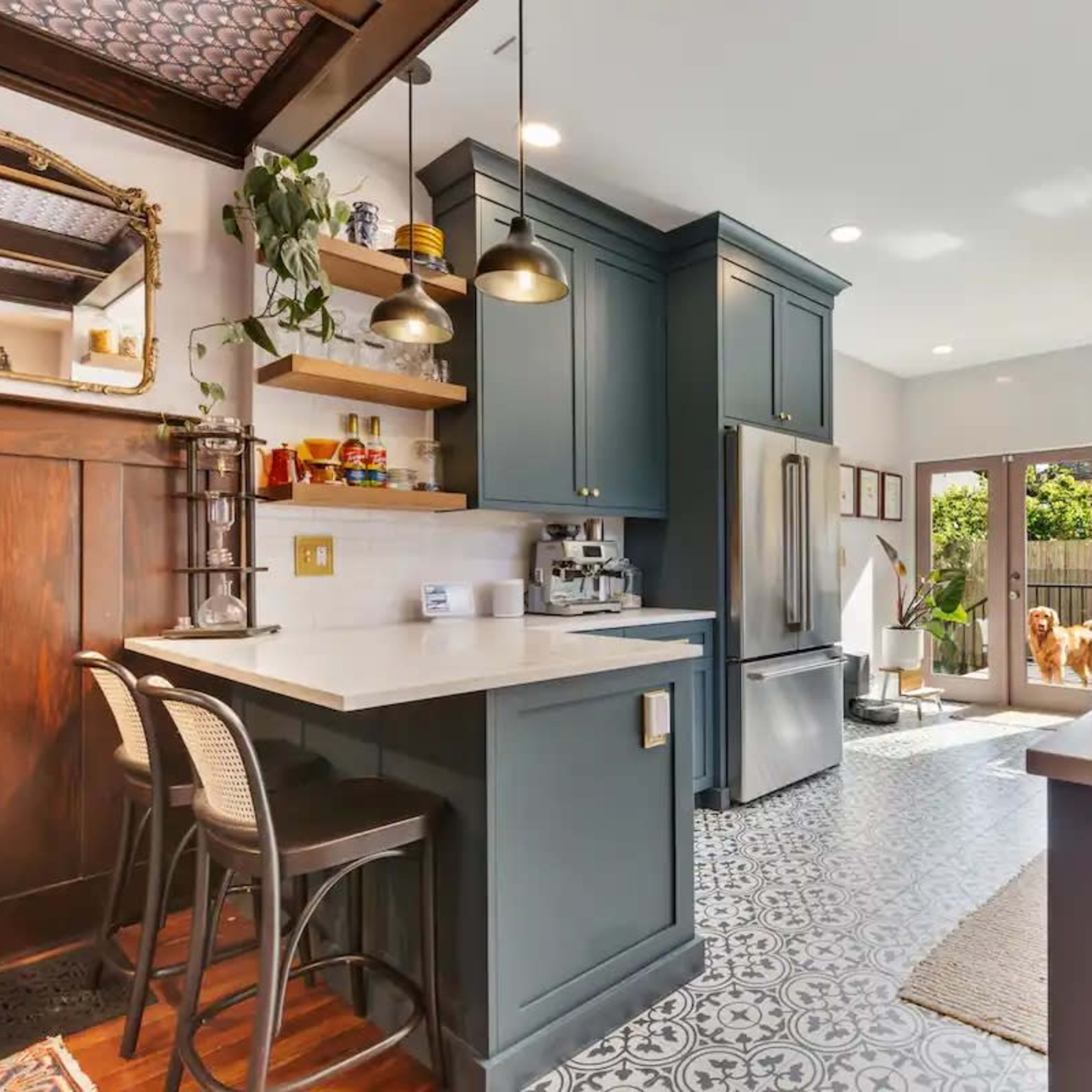 A modern kitchen features dark green cabinetry, white countertops, and patterned tile flooring, with an open door leading to a backyard.