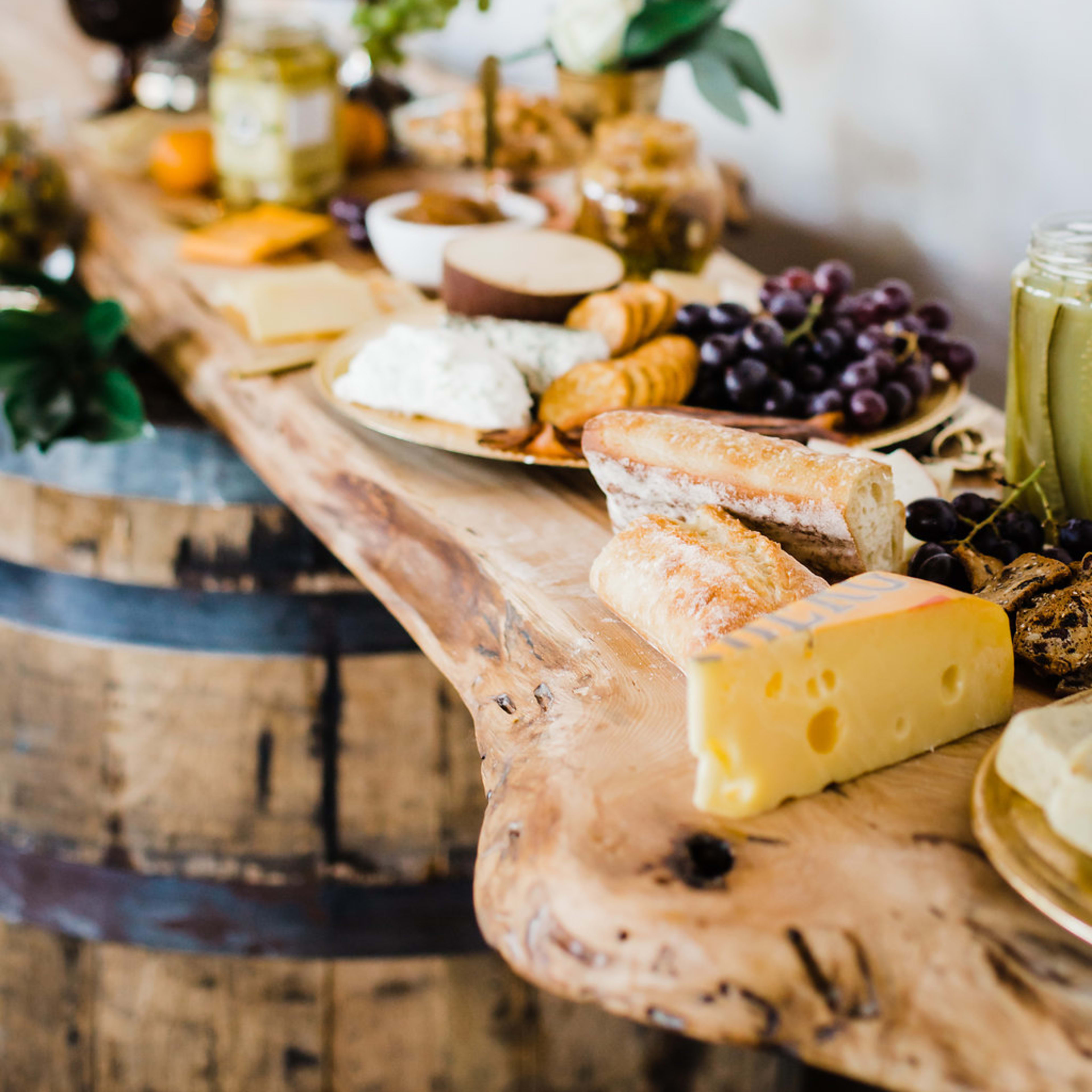 A long wooden table displays an assortment of cheeses, fruits, and spreads, arranged neatly for a gathering.