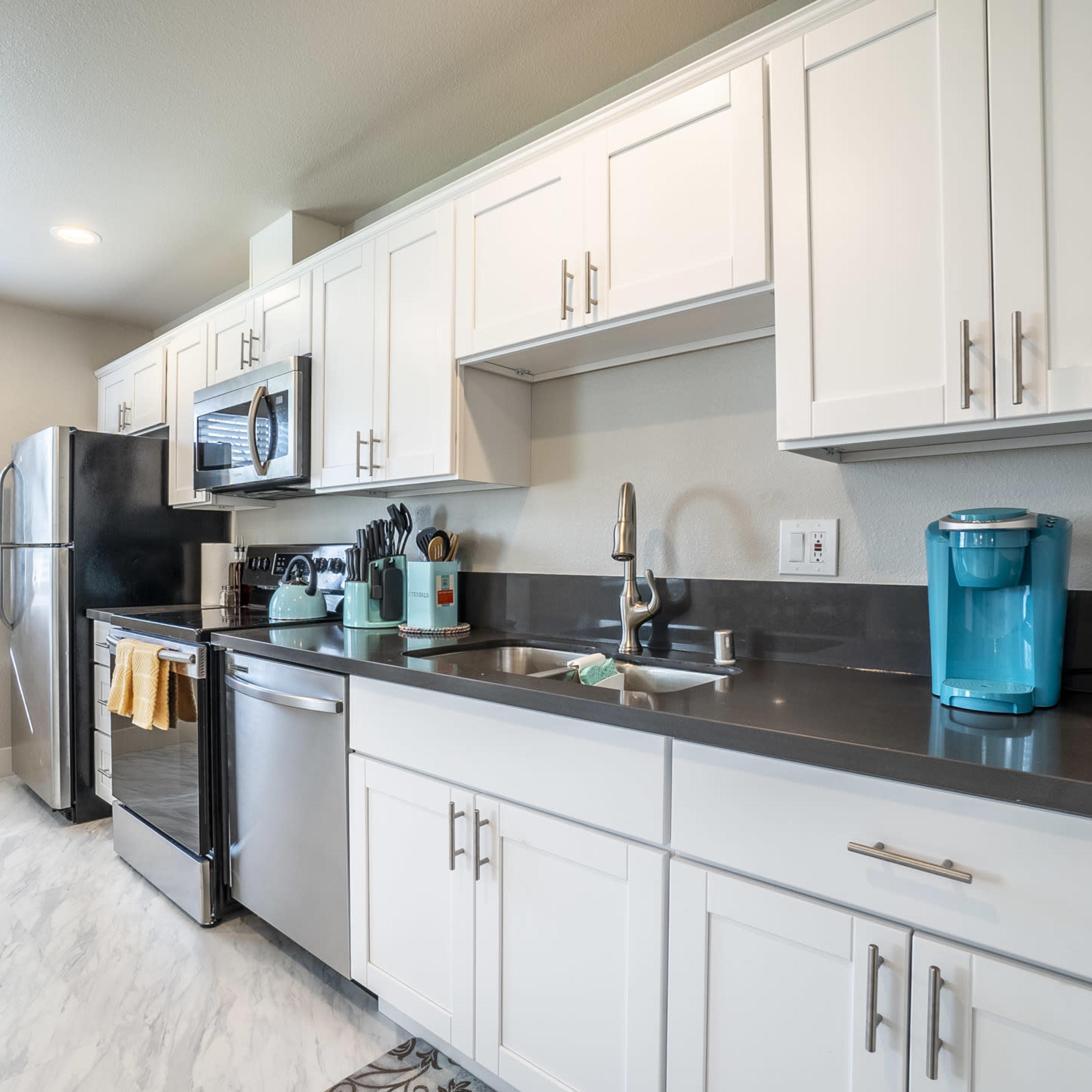 A modern kitchen features white cabinets, stainless steel appliances, and a blue countertop coffee maker beside a double sink.