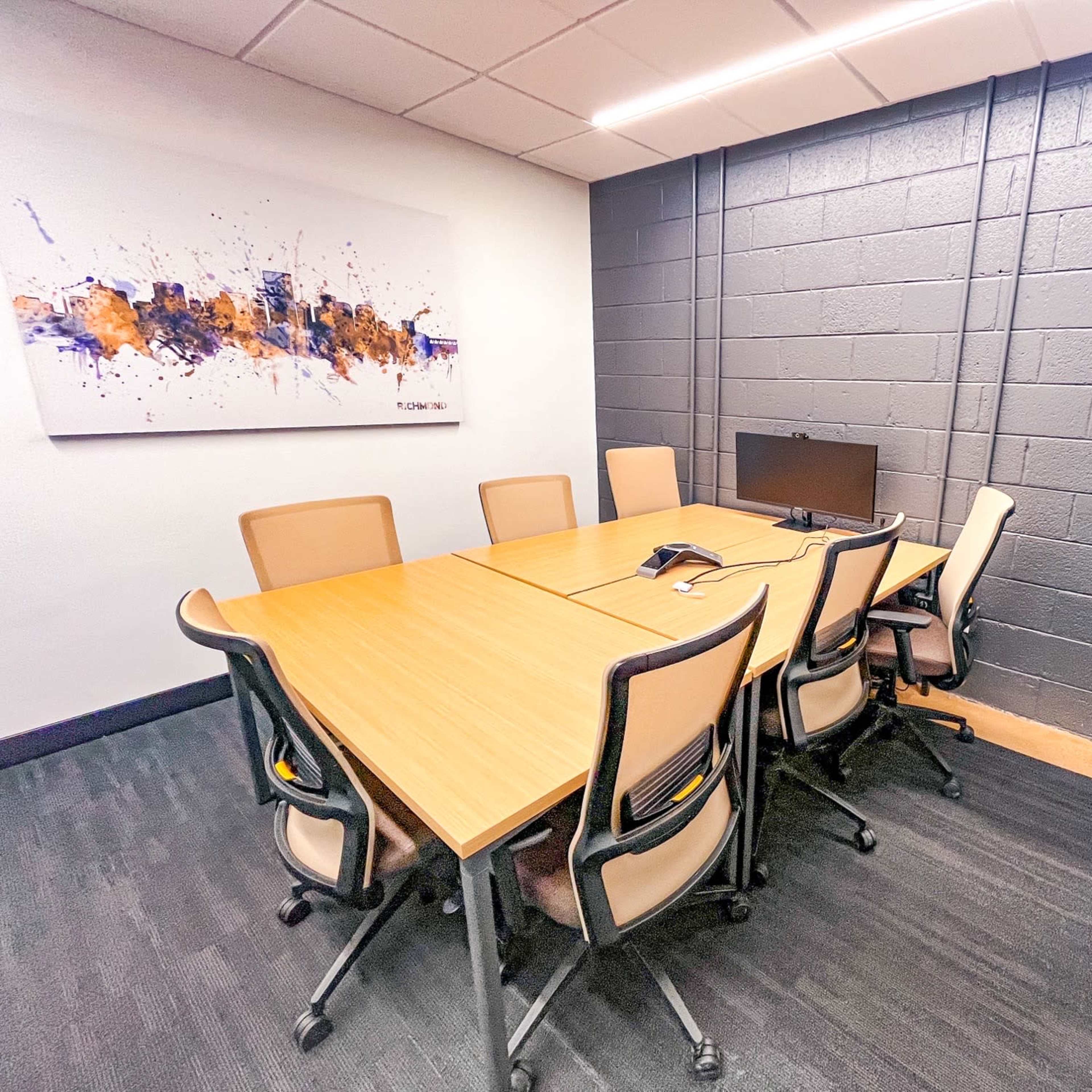 A conference room features a large wooden table surrounded by eight ergonomic chairs, with a monitor on one side and a potted plant in the corner.