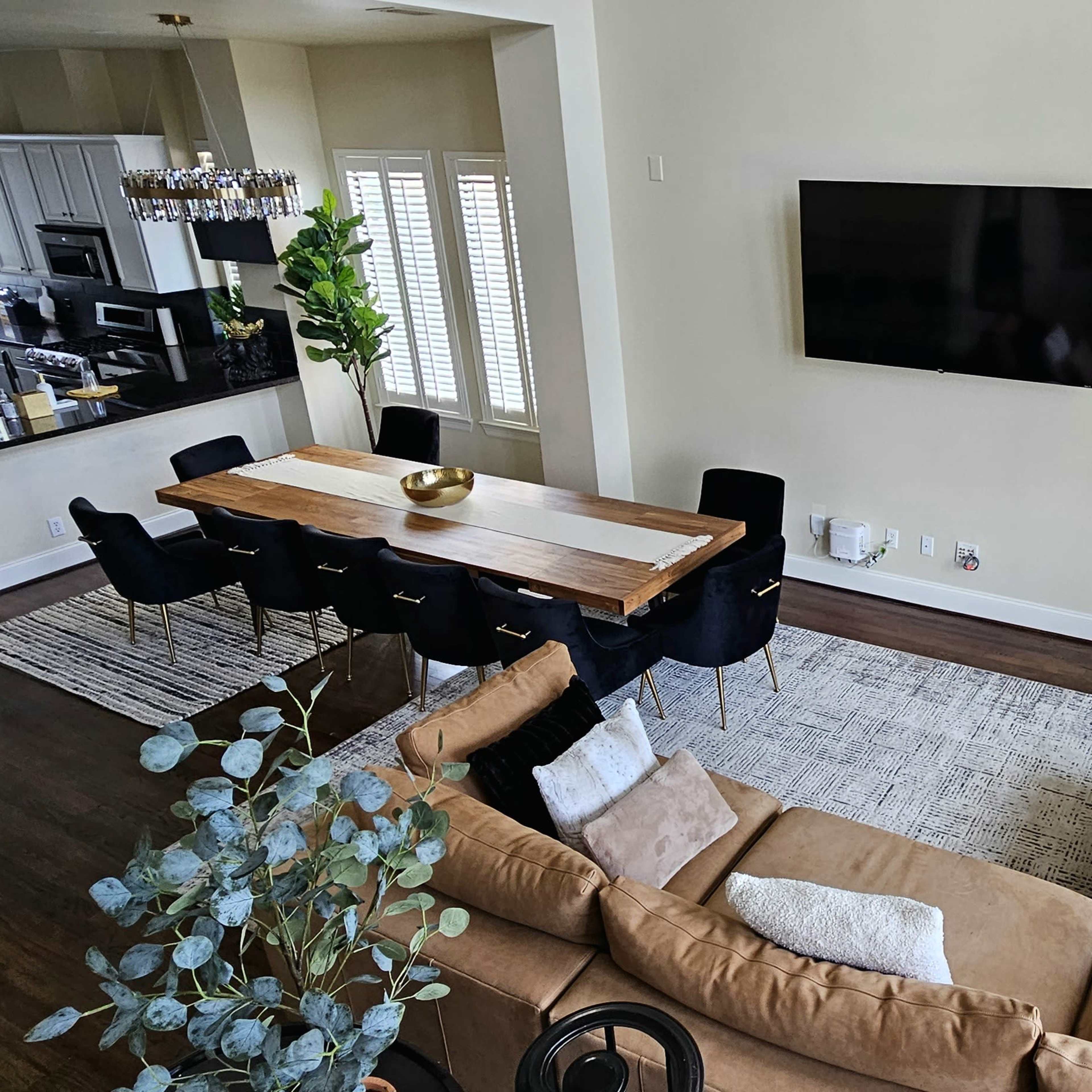 A spacious living area with a beige sofa, a wooden dining table surrounded by black chairs, and a large TV mounted on the wall.