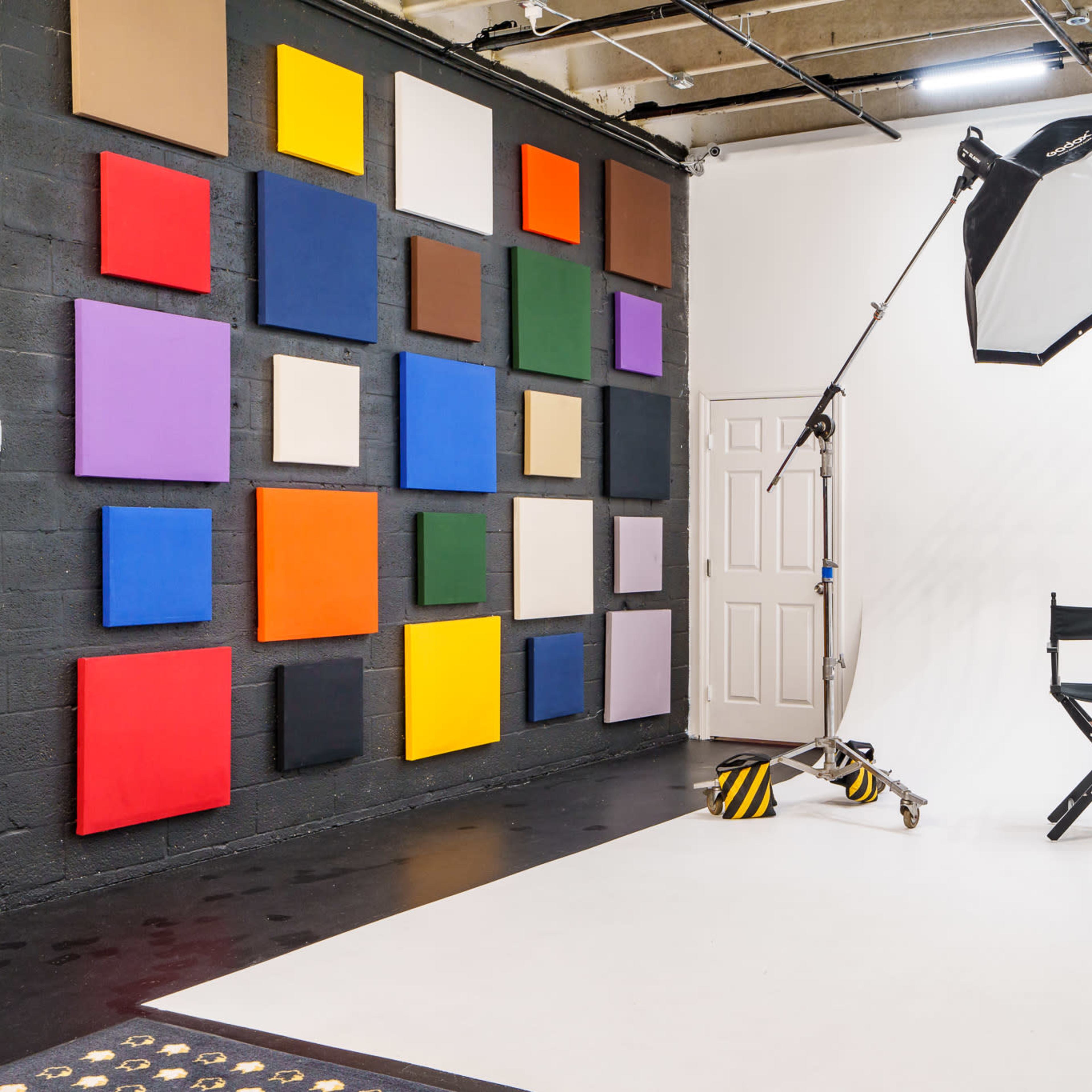 A photography studio featuring a wall of colorful canvases, a large lighting setup, and two director's chairs on a white backdrop.