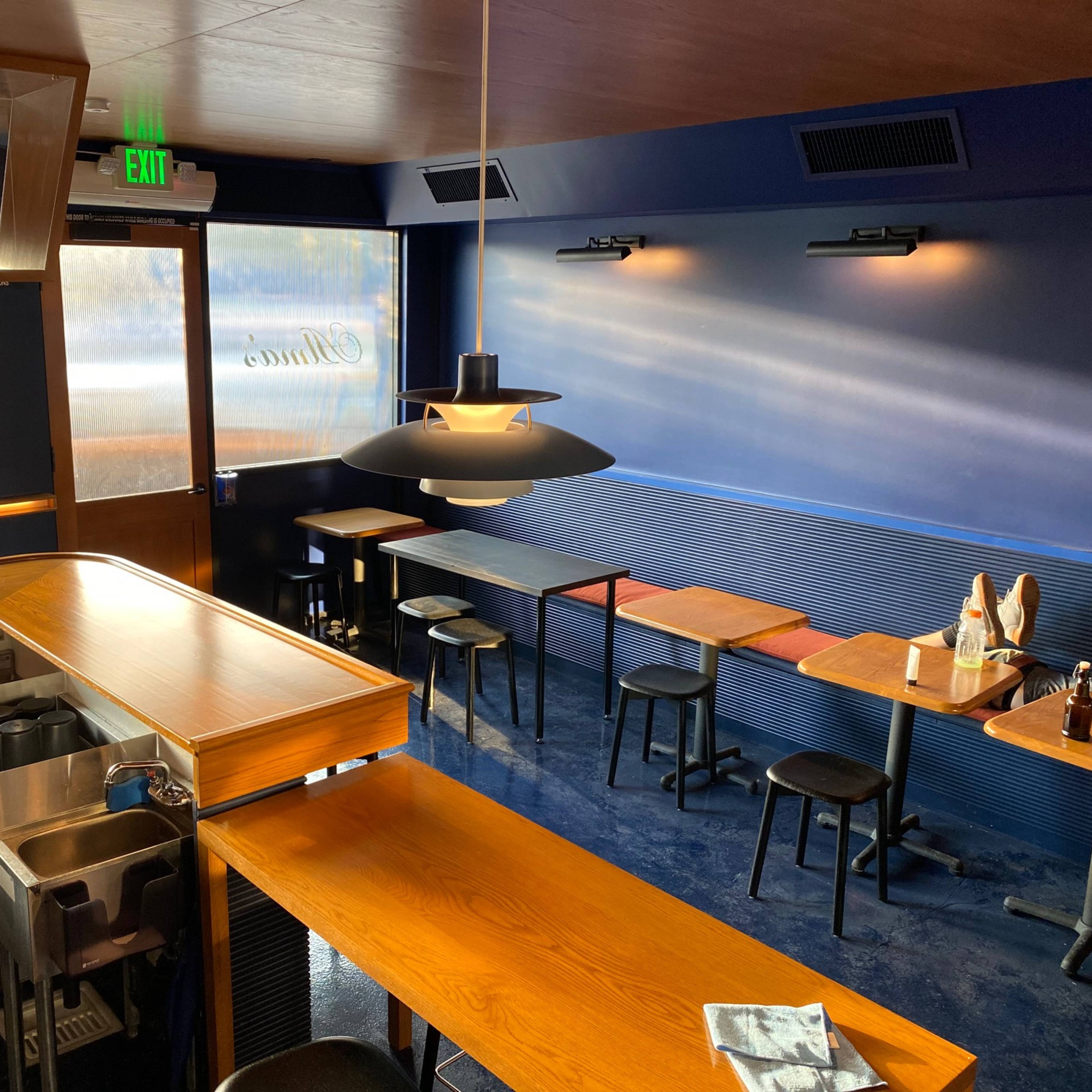 The interior of a modern restaurant with a wooden counter, black stools, and tables against a blue wall.