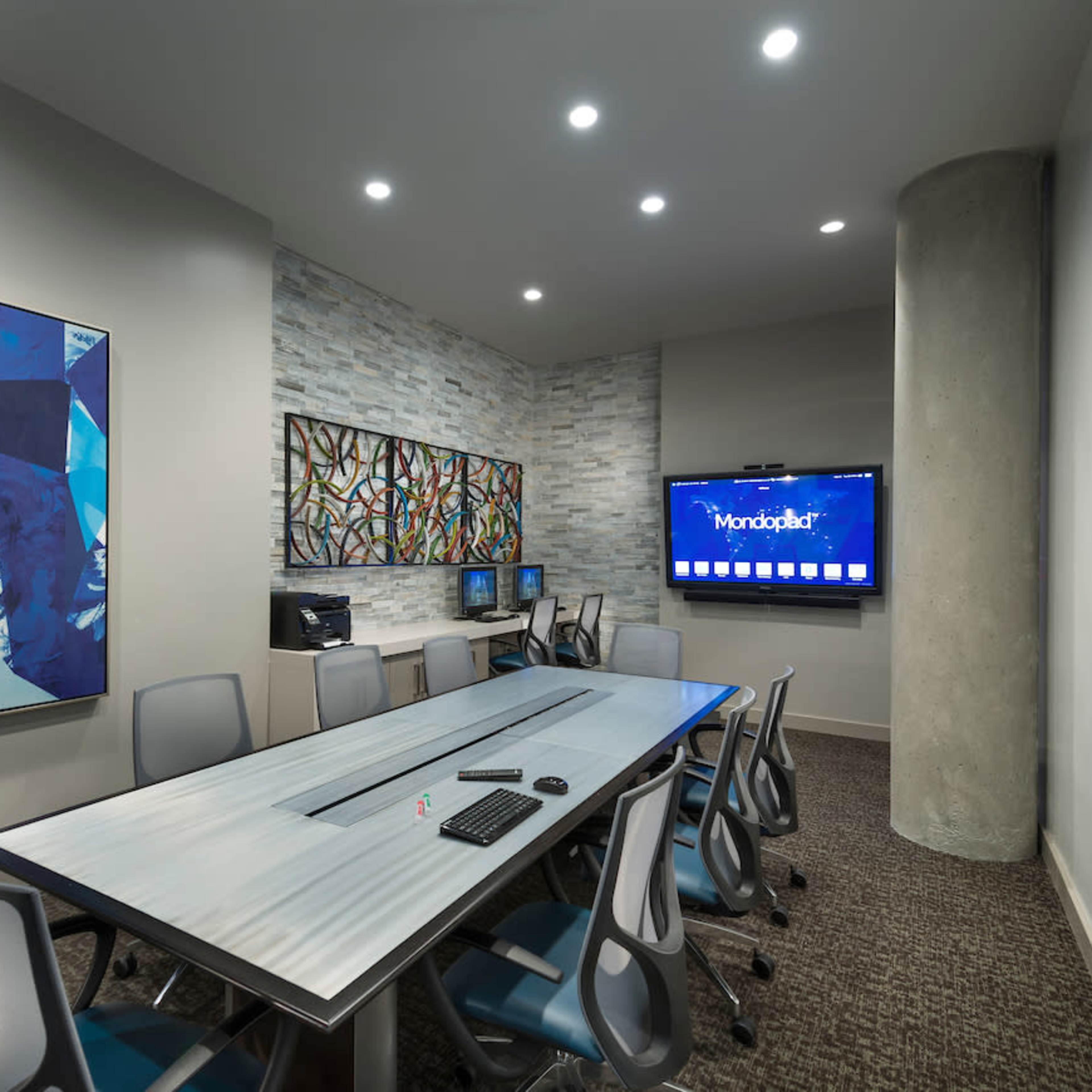 A modern conference room with a large table, several chairs, and two wall-mounted artworks, along with a television and computers in the background.