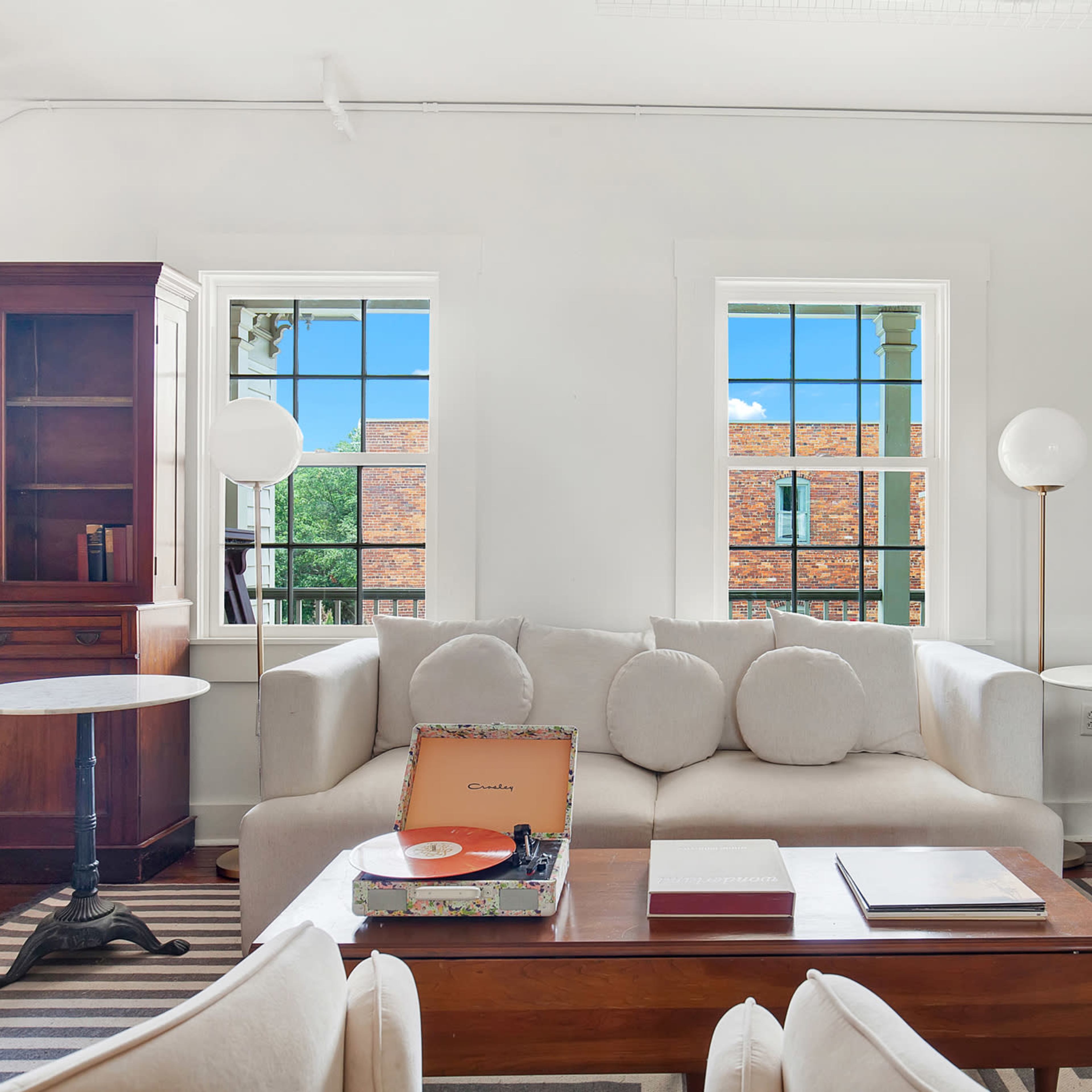 A modern living room with a beige sofa, two round side tables, a wooden coffee table with a vinyl record player, and large windows allowing natural light to enter.