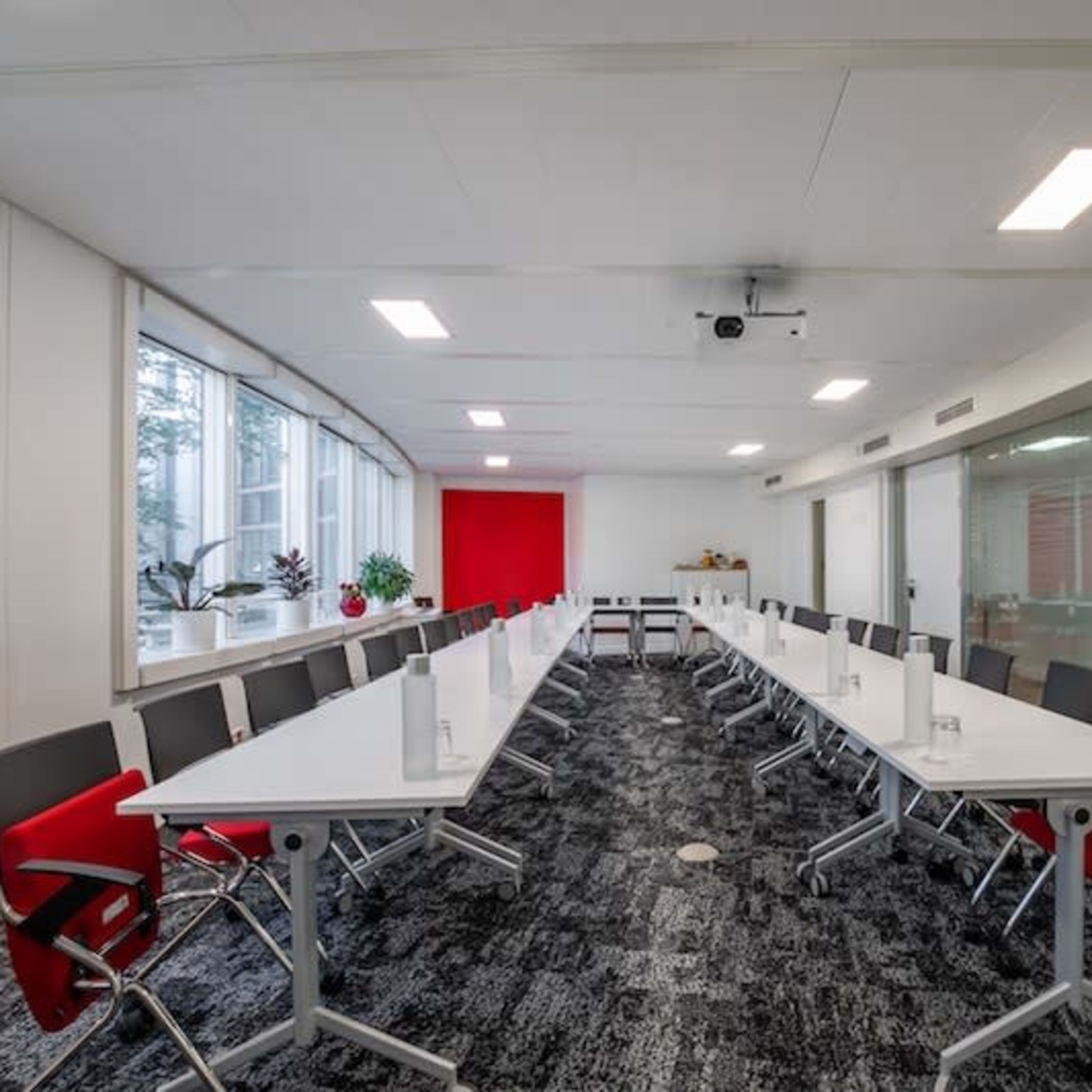 A spacious conference room features a long arrangement of tables and chairs, large windows, and a red accent wall.
