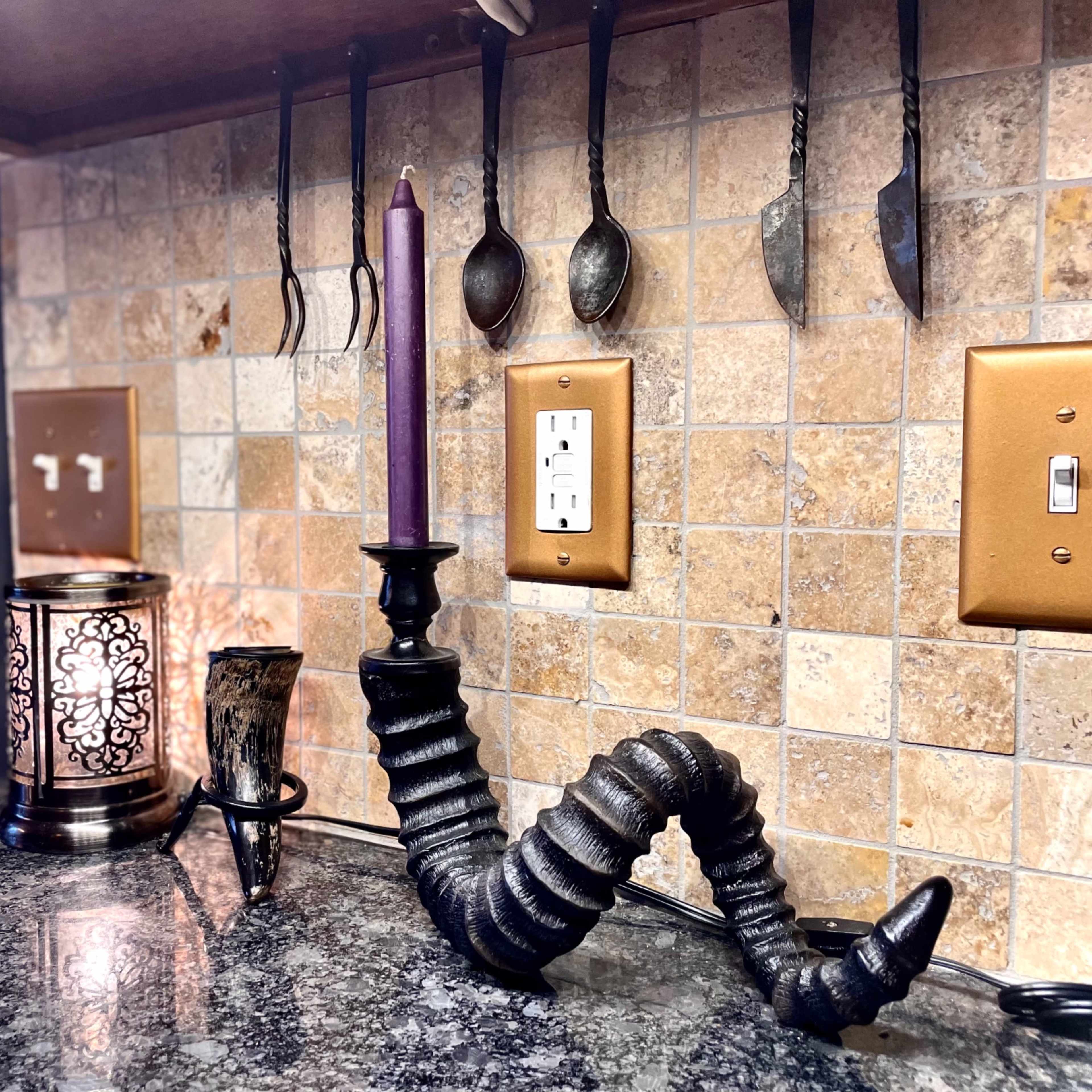 A dark, twisted candle holder with a purple candle, situated on a granite countertop alongside a decorative lantern and a silver spoon holder against a tiled backsplash.