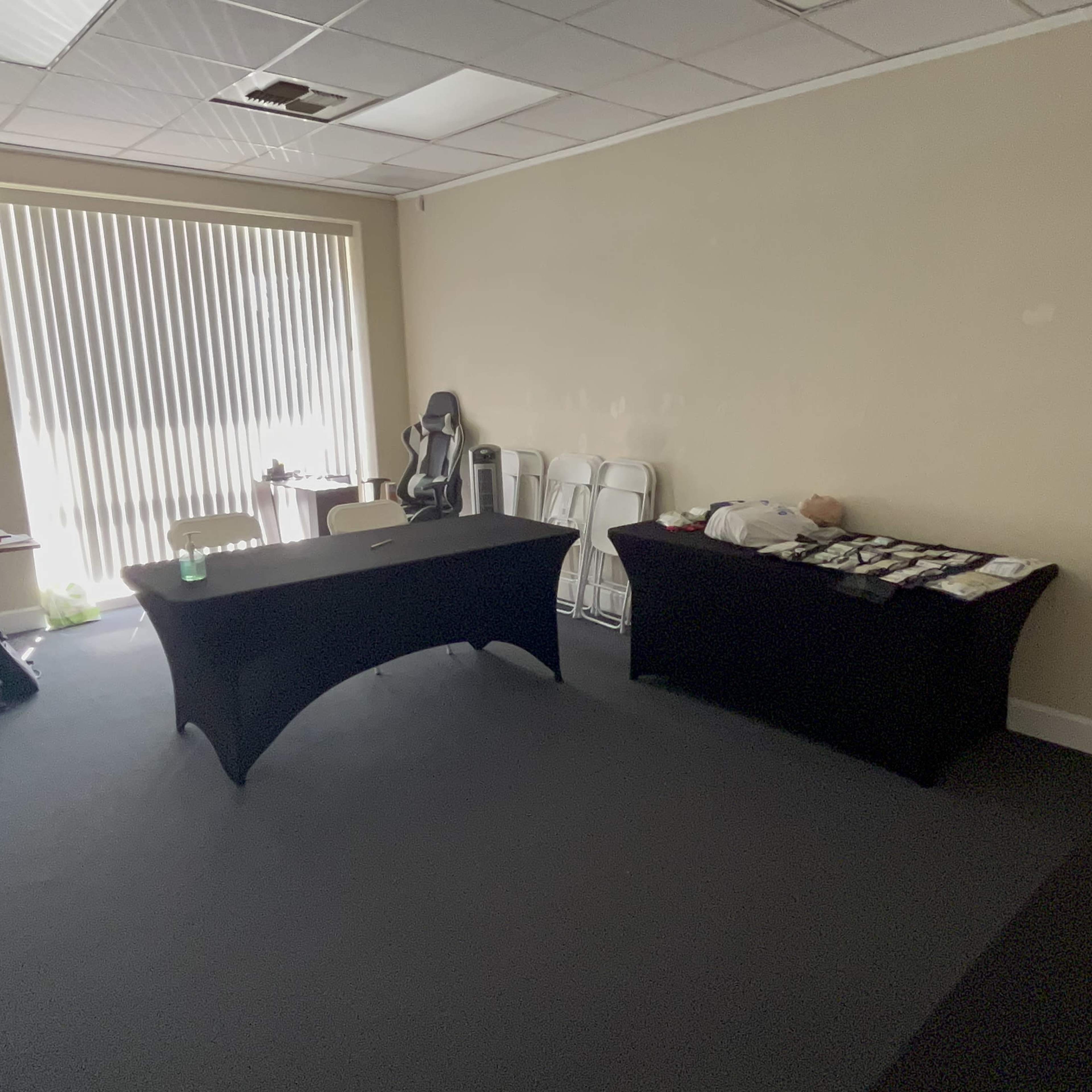 A sparsely furnished room with two tables covered in black cloth and a row of white chairs against one wall, illuminated by natural light from a large window.