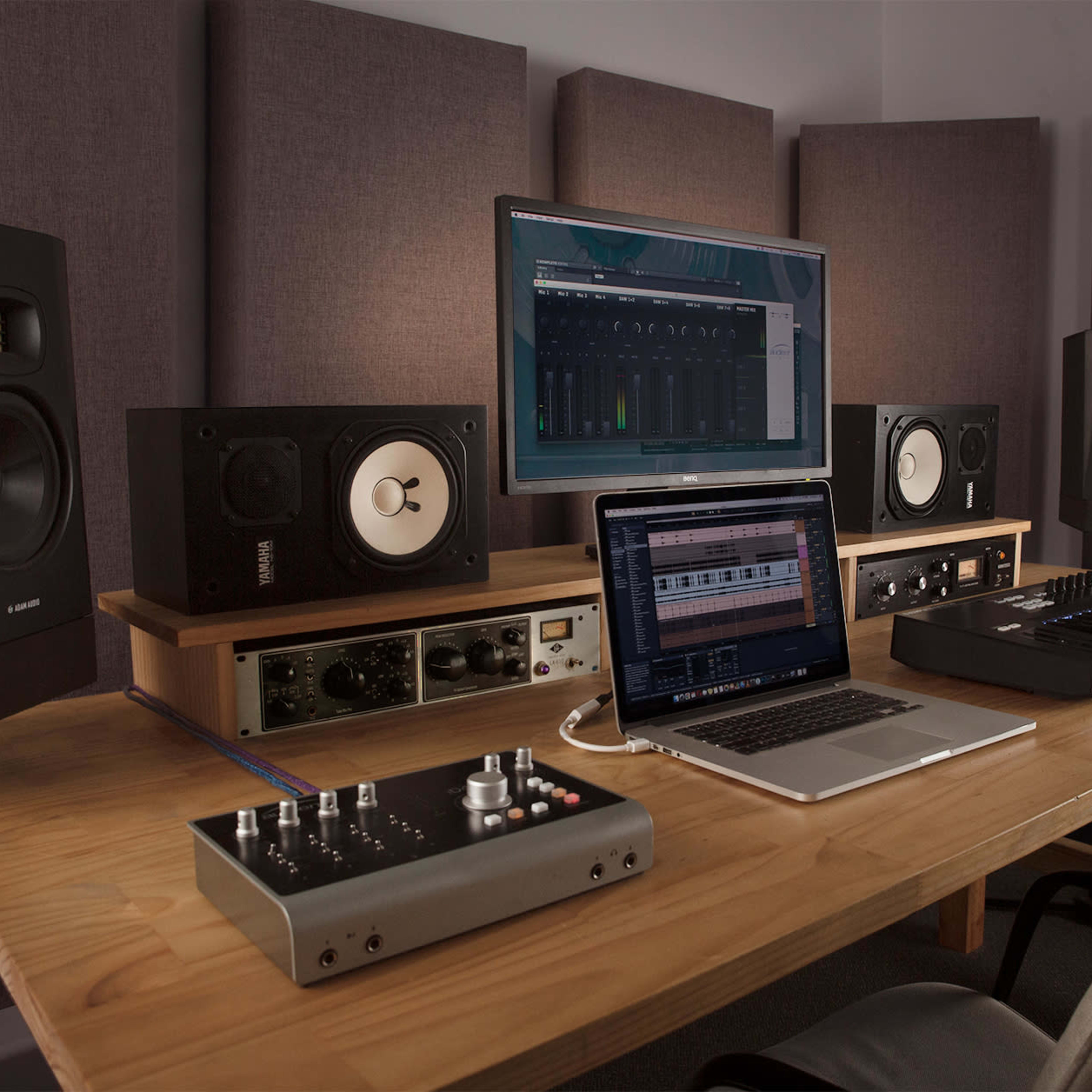A wooden desk in a music studio features a laptop, audio interface, MIDI keyboard, and two studio monitors, with acoustic panels on the wall behind.