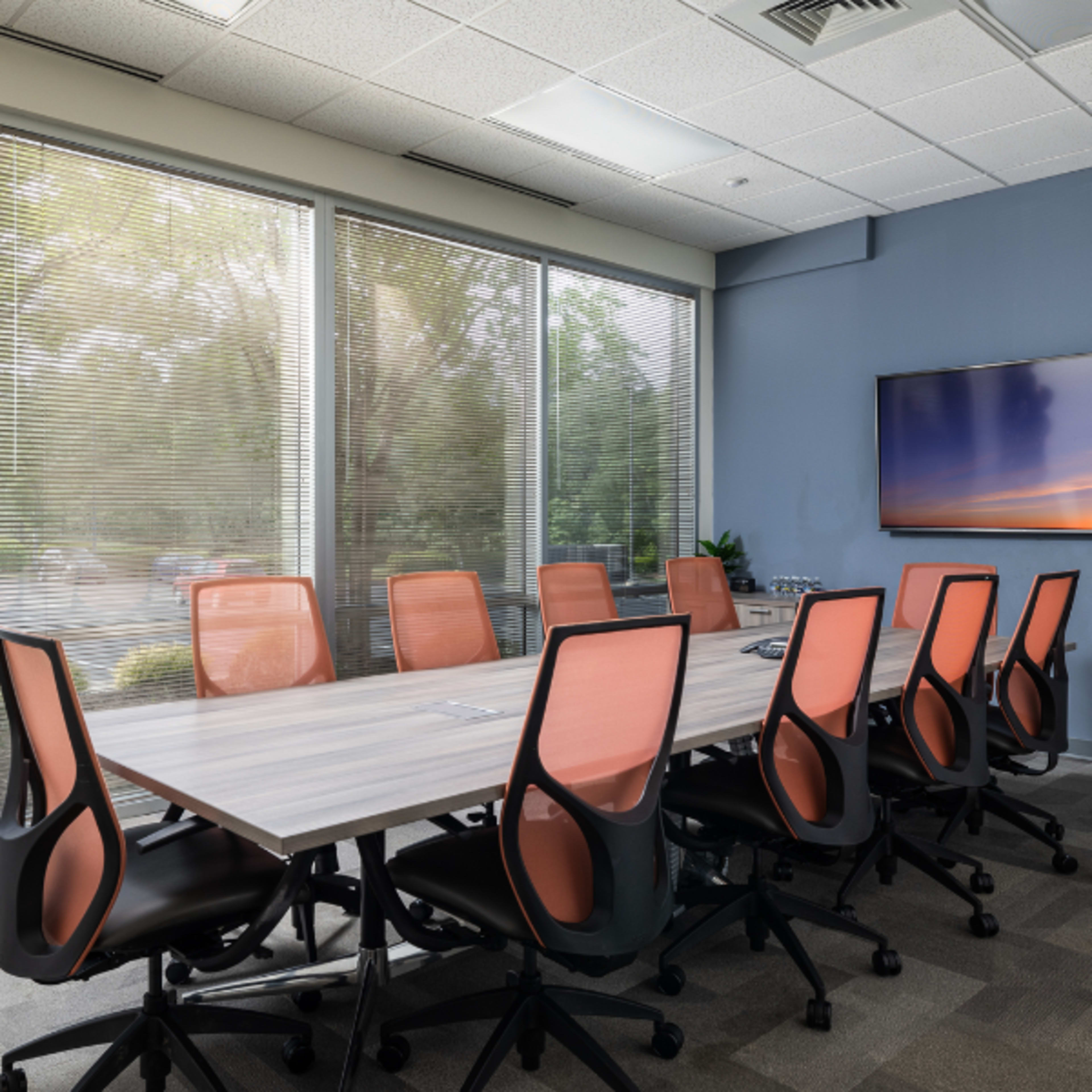 A modern conference room features a long table surrounded by eight ergonomic chairs, with large windows allowing natural light and a television mounted on a blue wall.