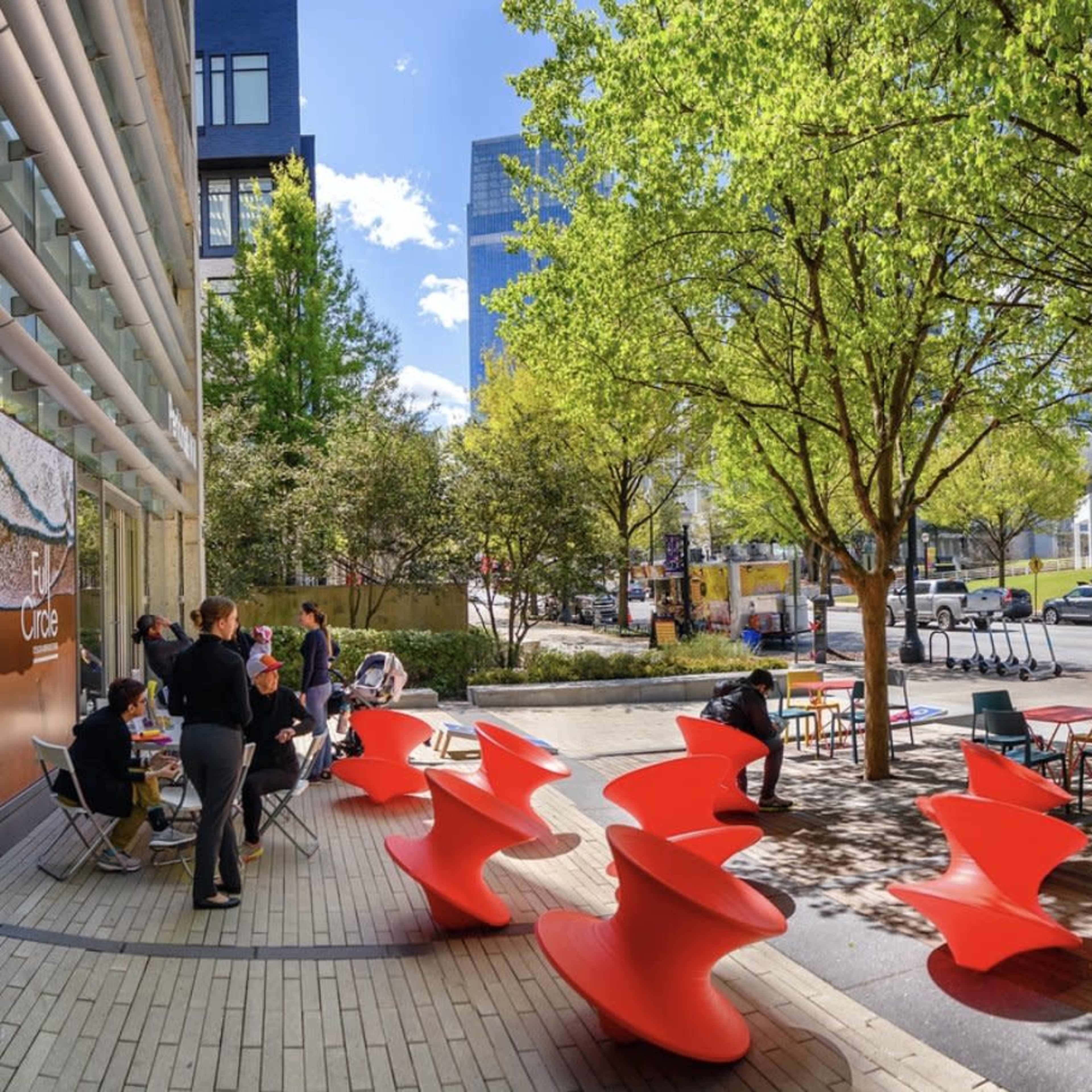 A modern outdoor space with colorful red seating, trees, and people enjoying the area near a building and street.