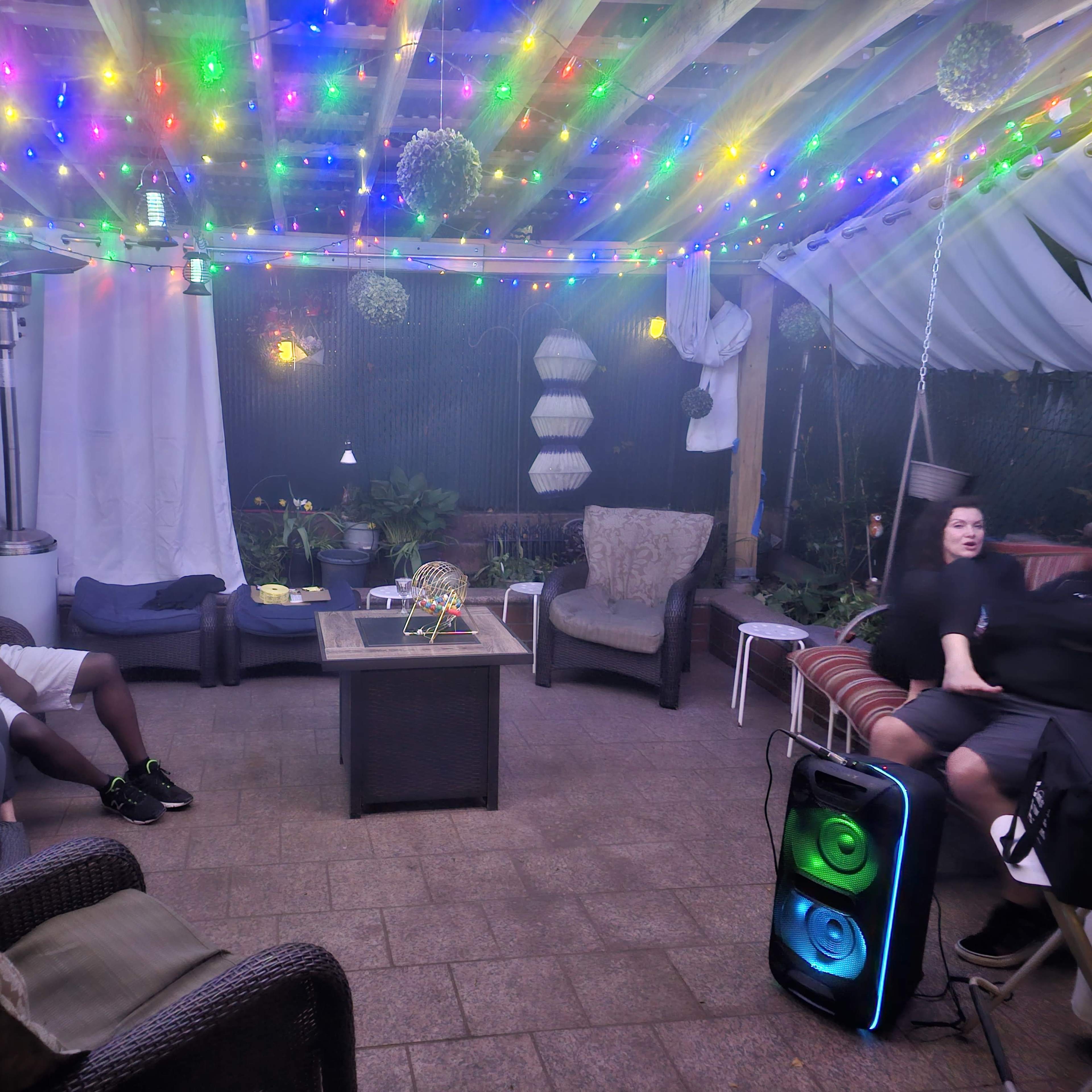 A cozy outdoor space features several people sitting on comfortable furniture under colorful string lights.
