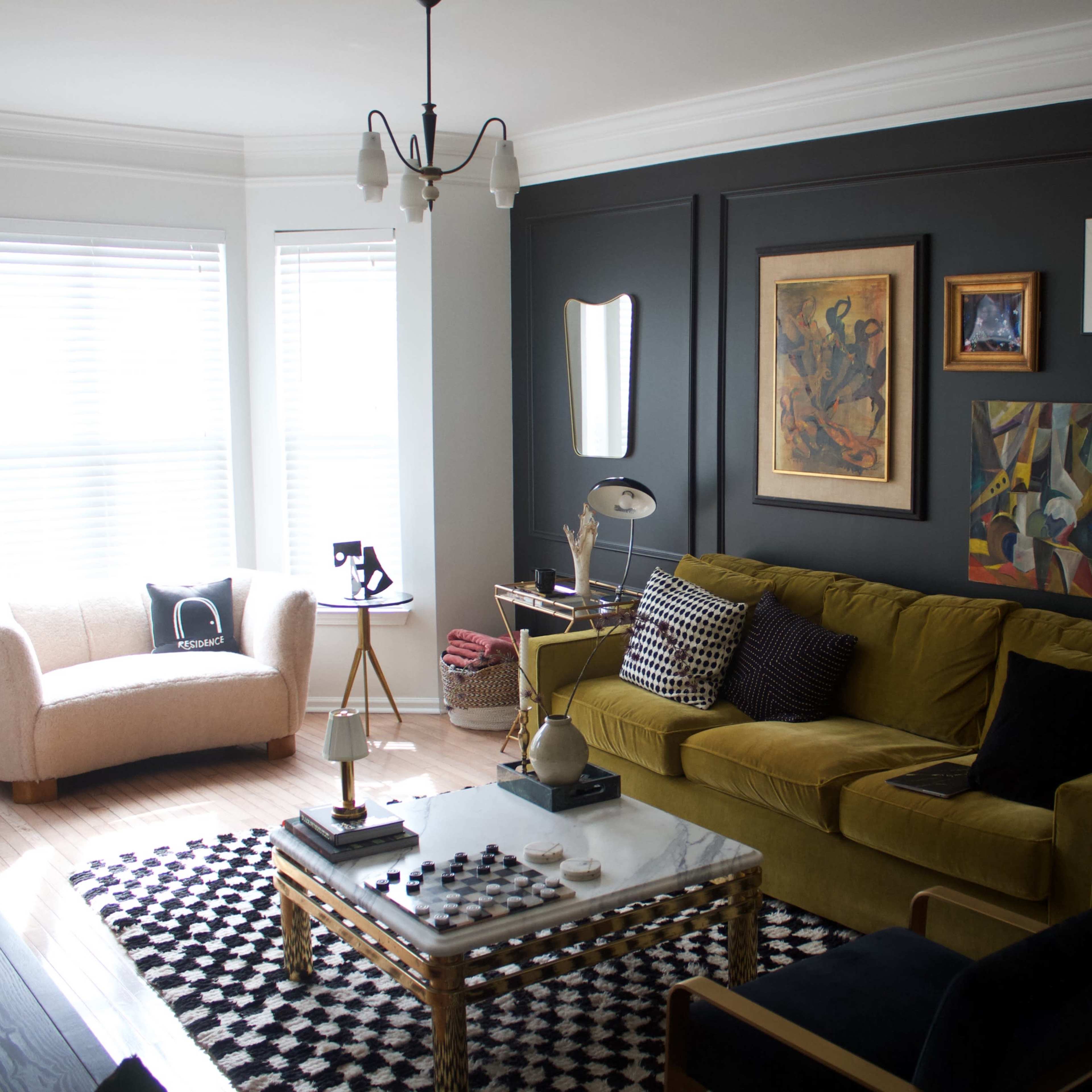A living room featuring a green sofa, a coffee table, artwork on the walls, and large windows with white blinds.