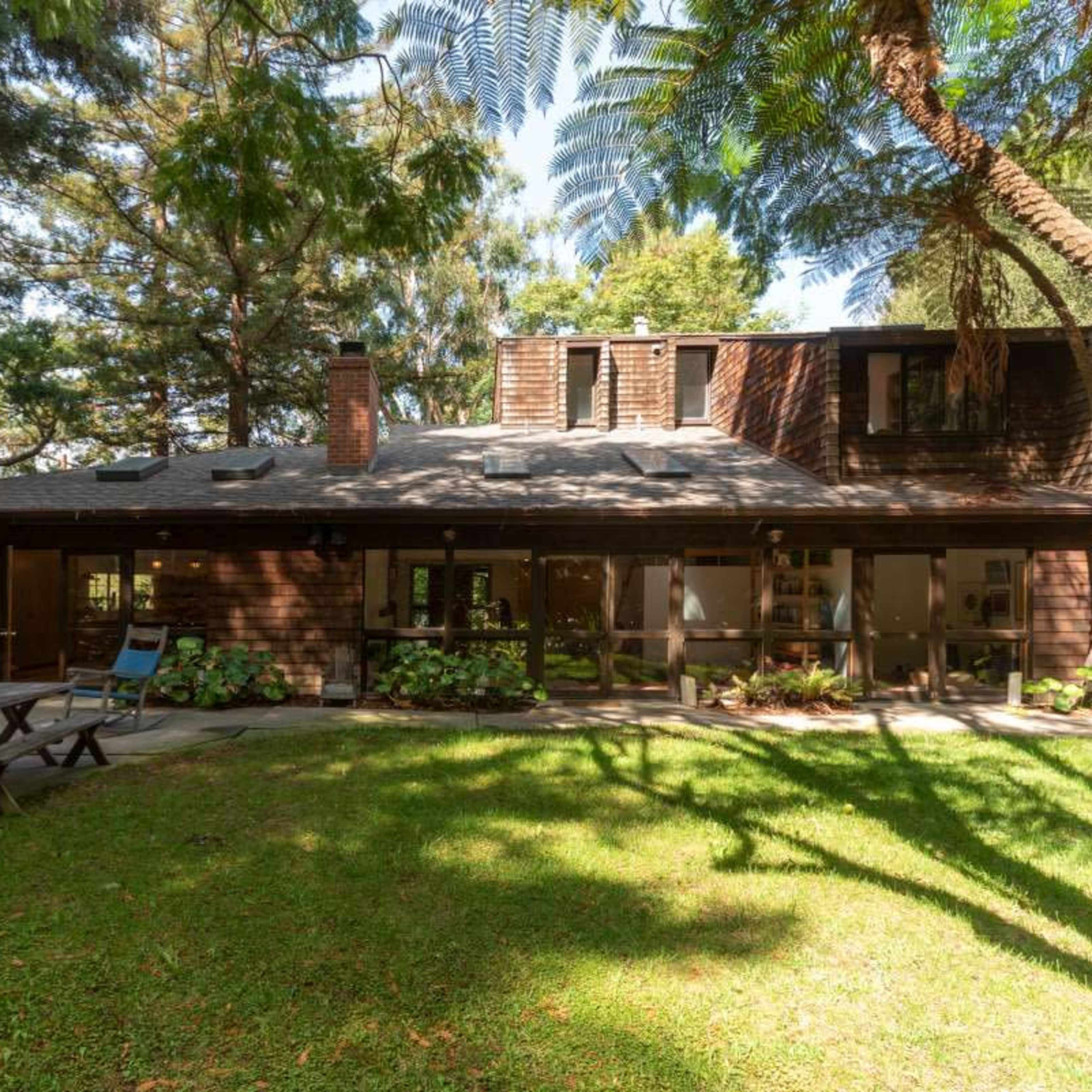 A wooden house with a sloped roof, surrounded by trees and a grassy area, features a patio and a picnic table in the backyard.