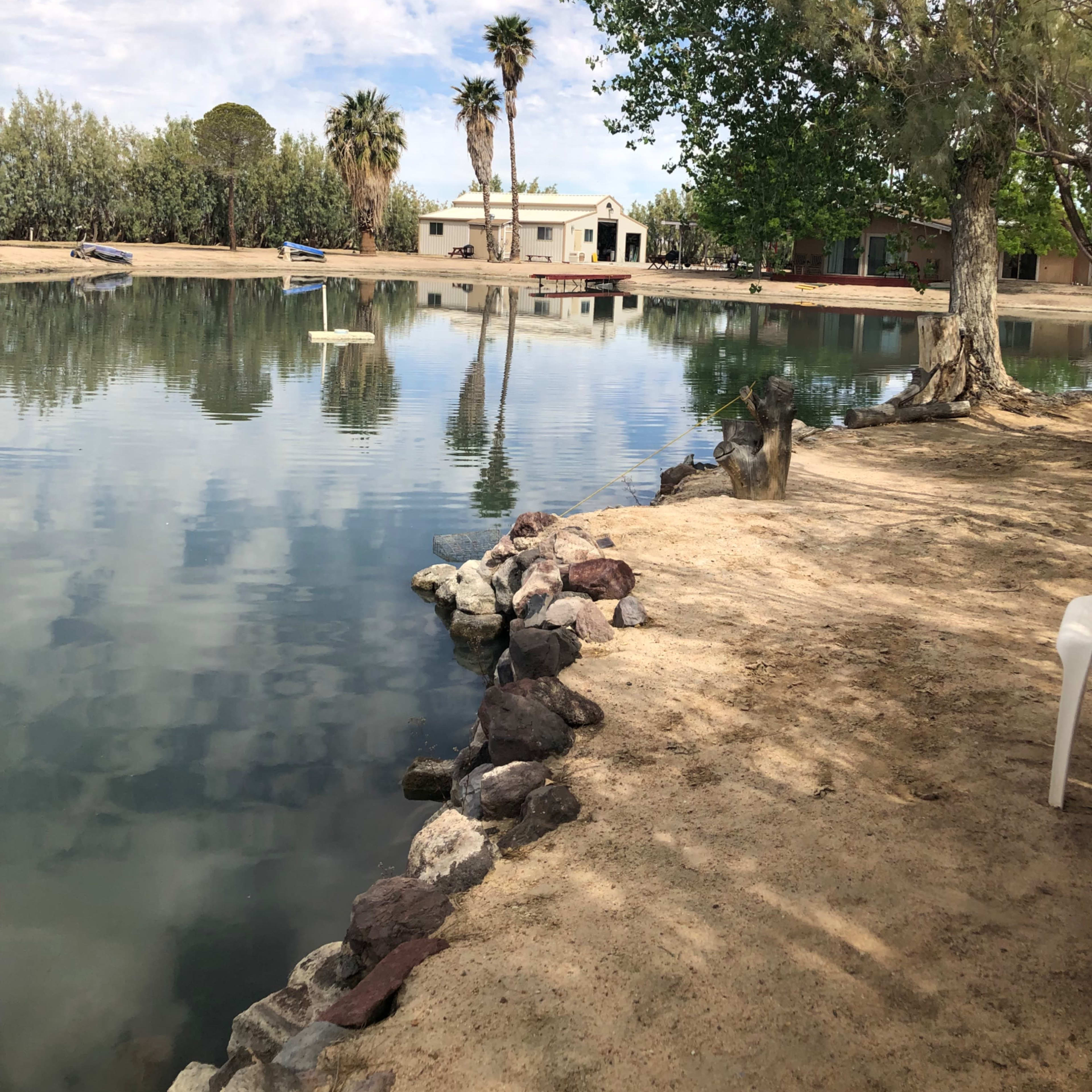 A tranquil lake bordered by sandy soil and rocks, with palm trees and a residential building visible in the background under a partly cloudy sky.
