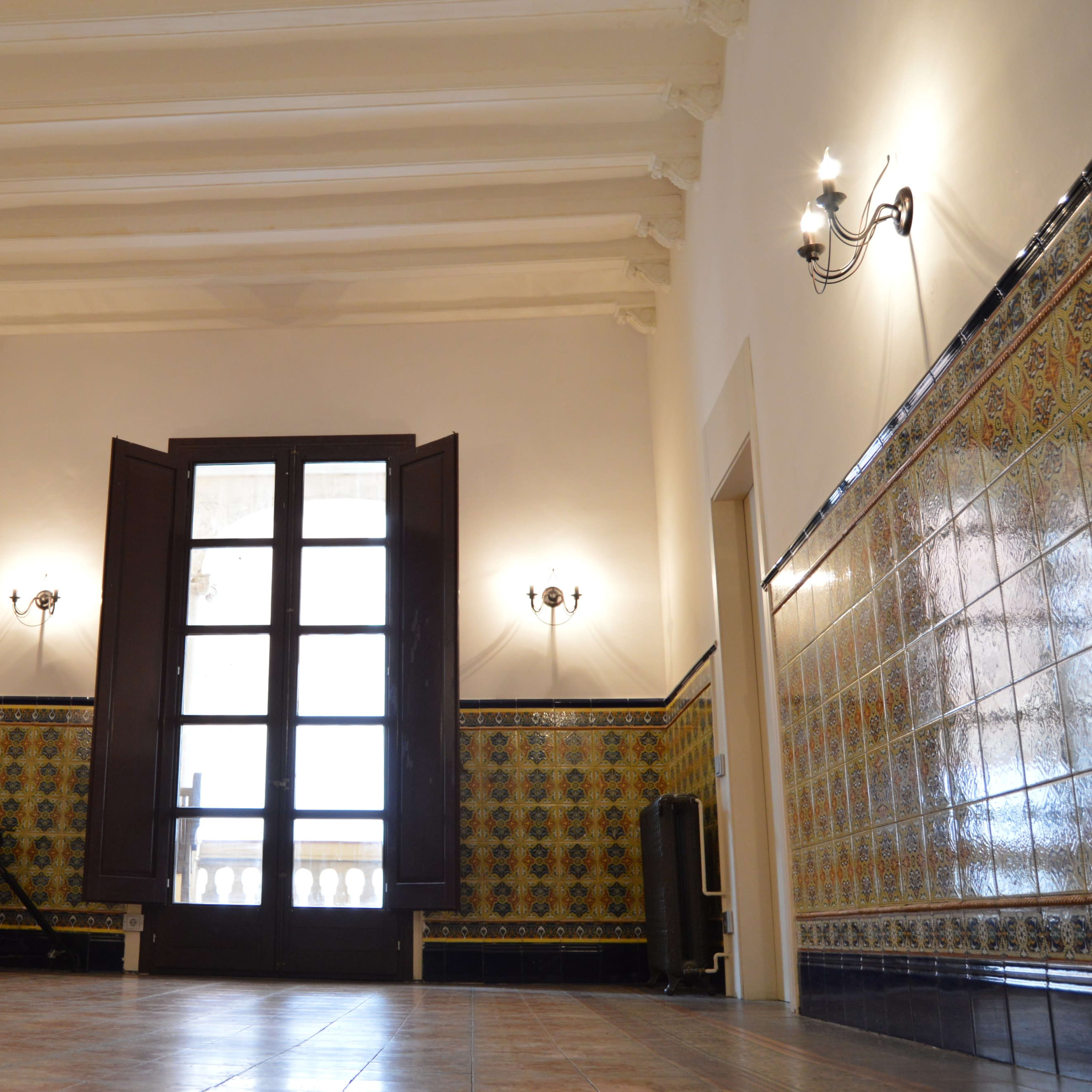 A spacious interior room with patterned tile walls, a set of open wooden doors leading to a balcony, and exposed ceiling beams.