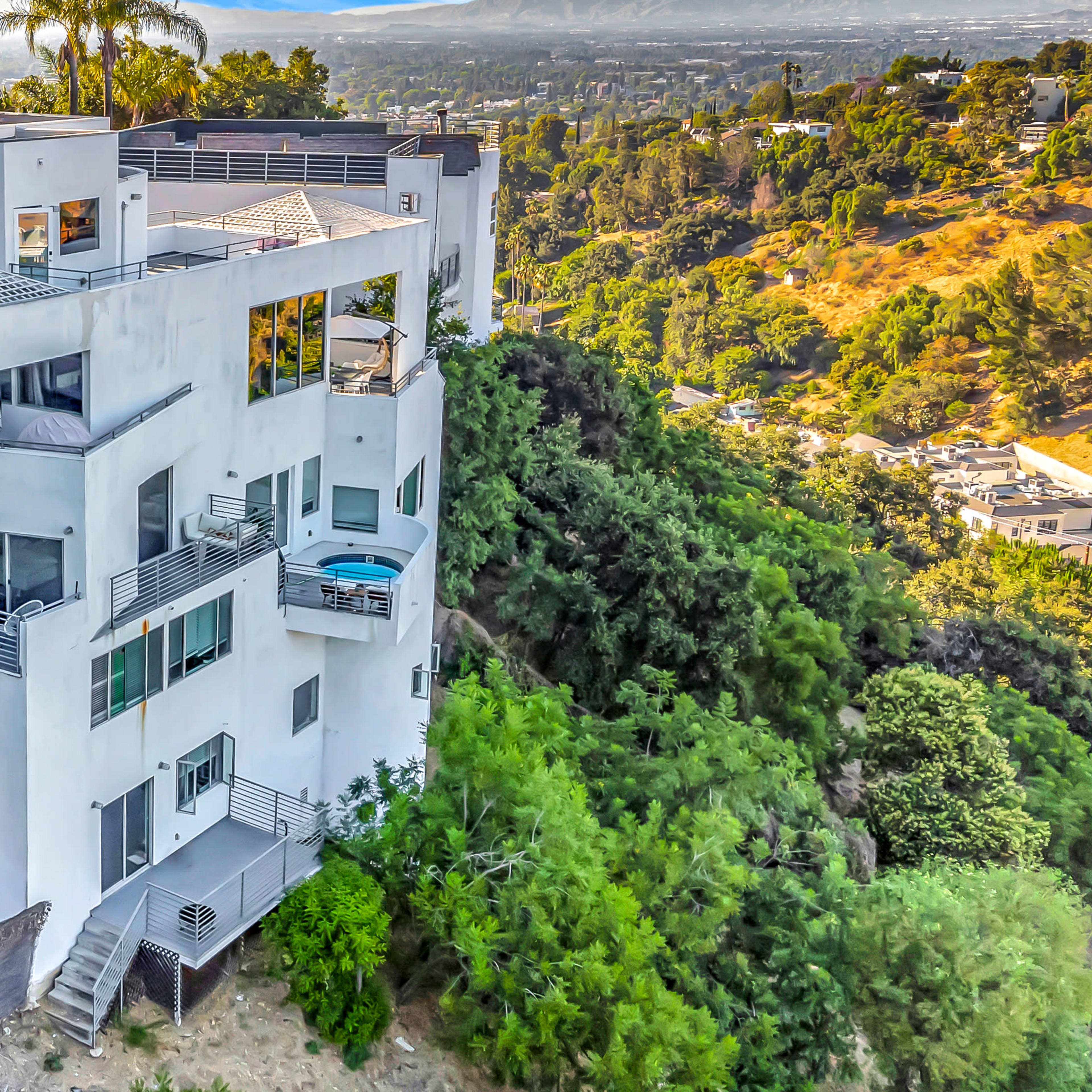 A modern multi-level white house is situated on a hillside, surrounded by greenery and featuring multiple balconies and staircases.