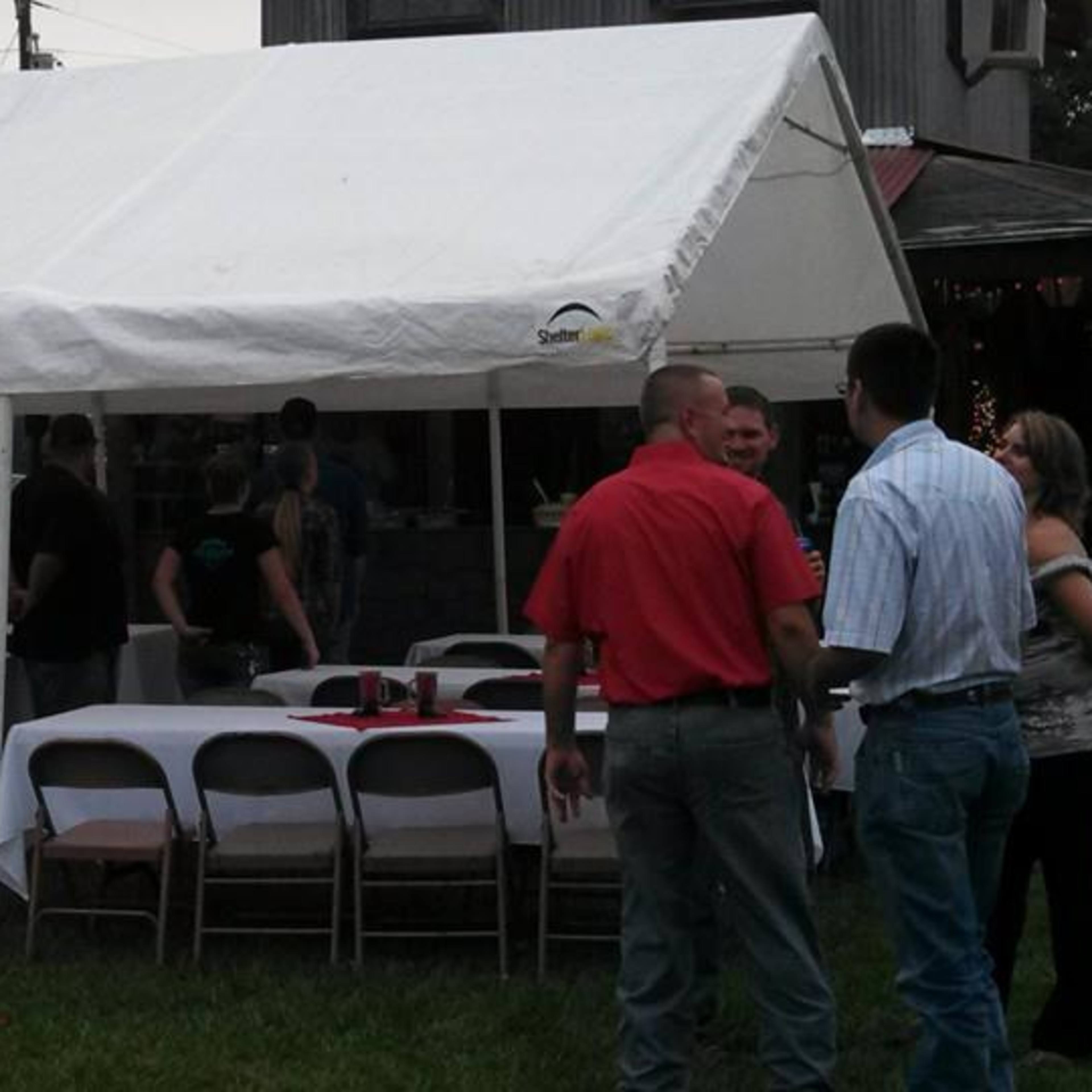 A group of people gathers around tables under a white tent in a grassy area, with a building visible in the background.