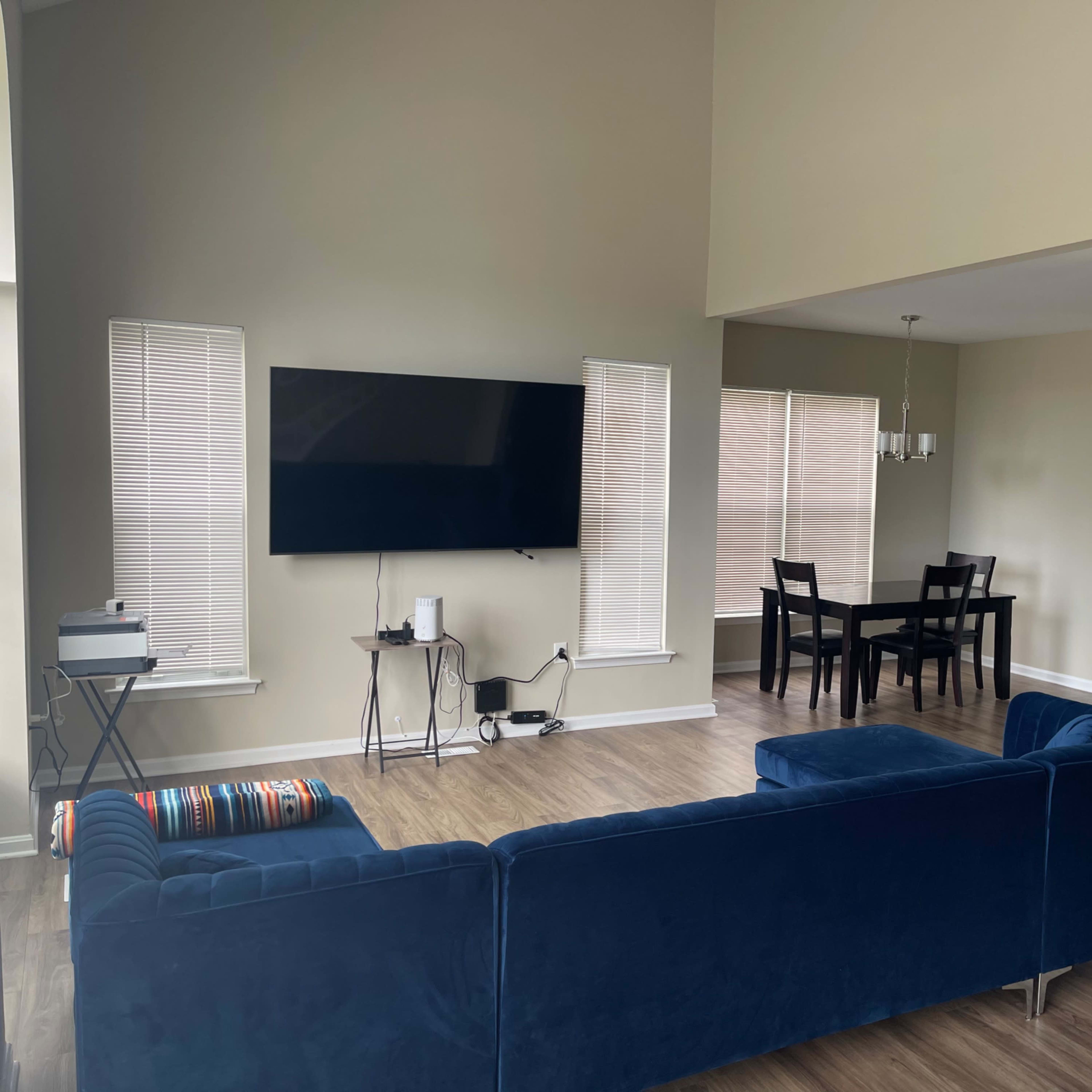 A spacious living area with a blue sectional sofa, a mounted television on the wall, a dining table with black chairs, and large windows with blinds.