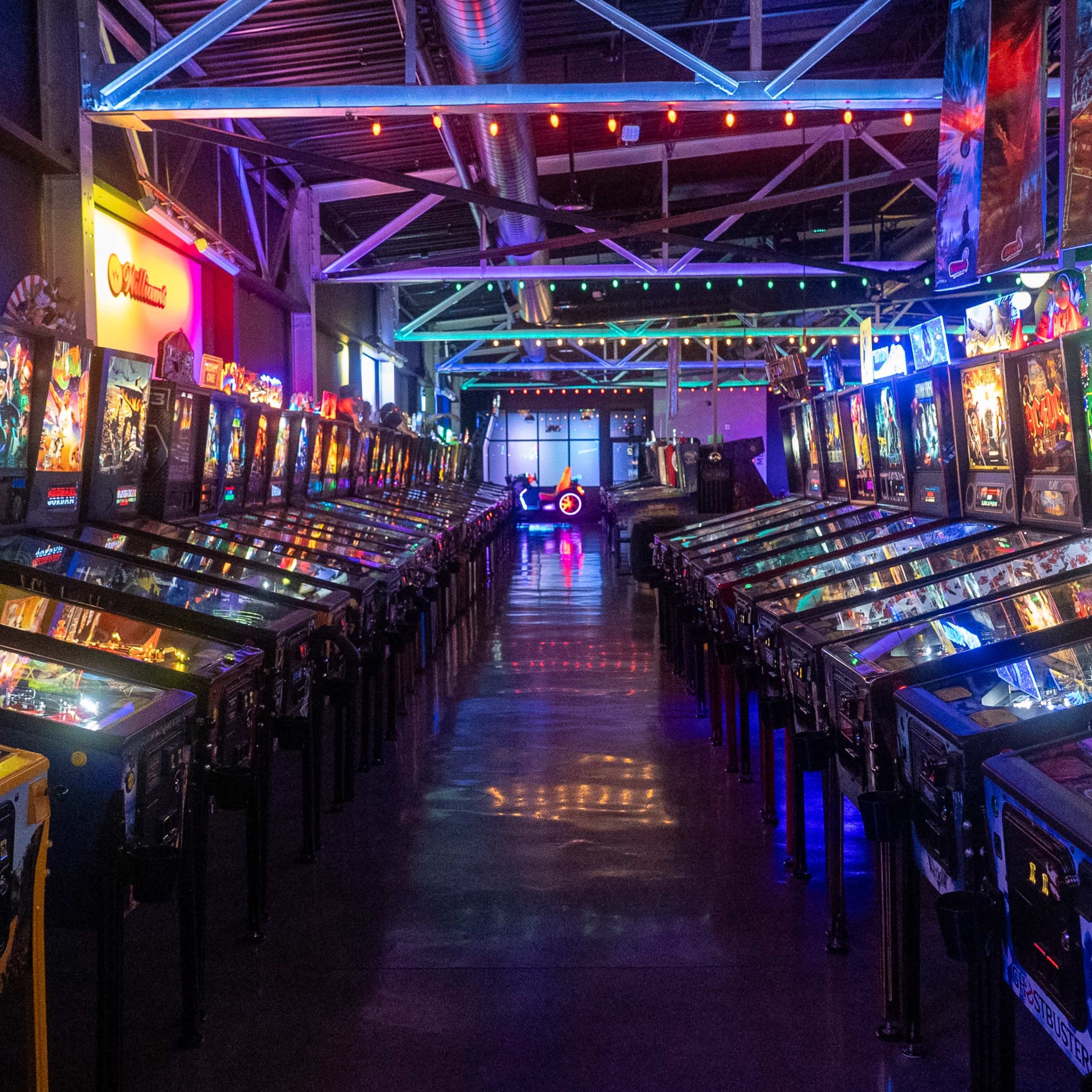 A row of brightly lit pinball machines lines the floor of a spacious arcade, with colorful neon lights reflecting off the glass surfaces.