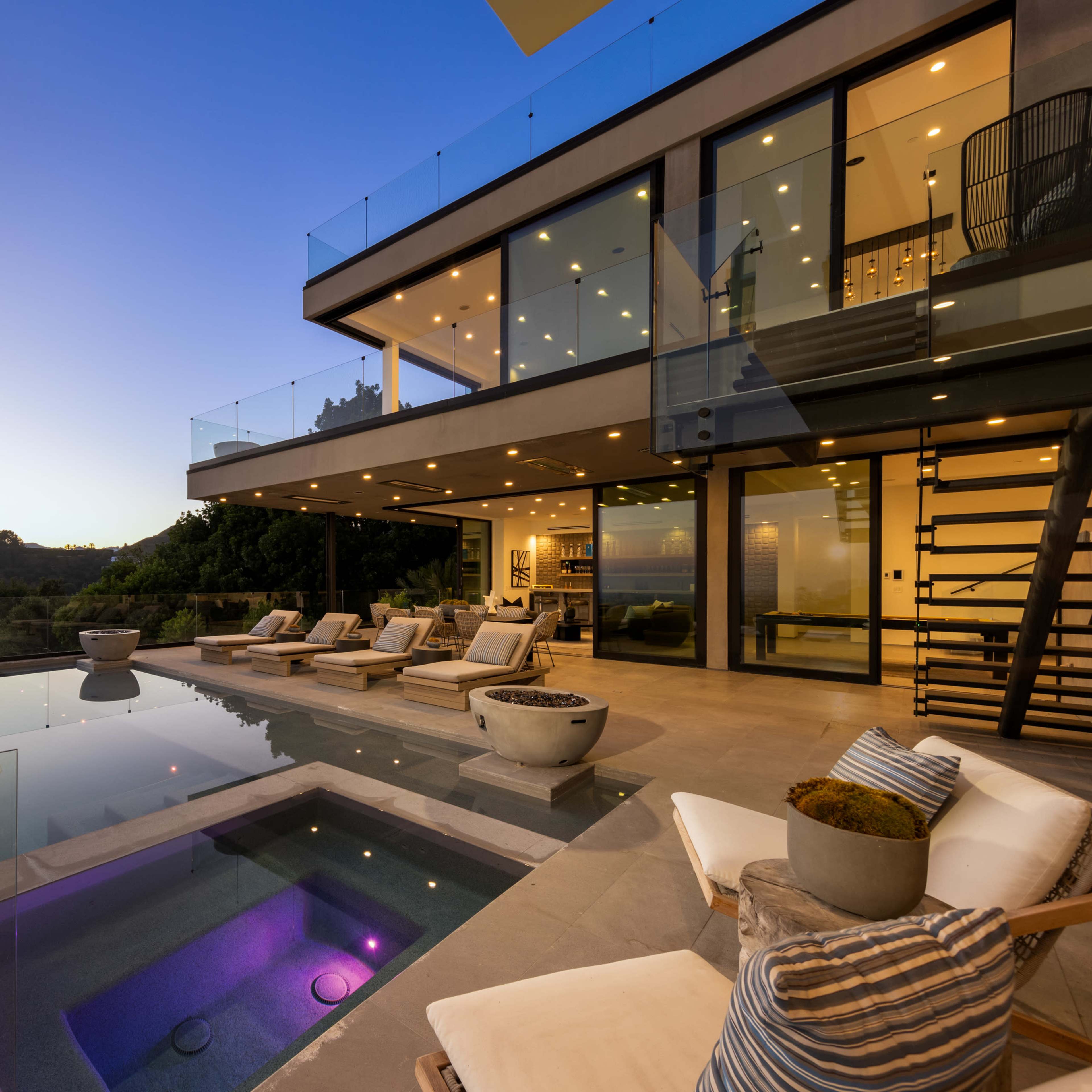 A modern house with large glass walls overlooks a swimming pool and spa at dusk.
