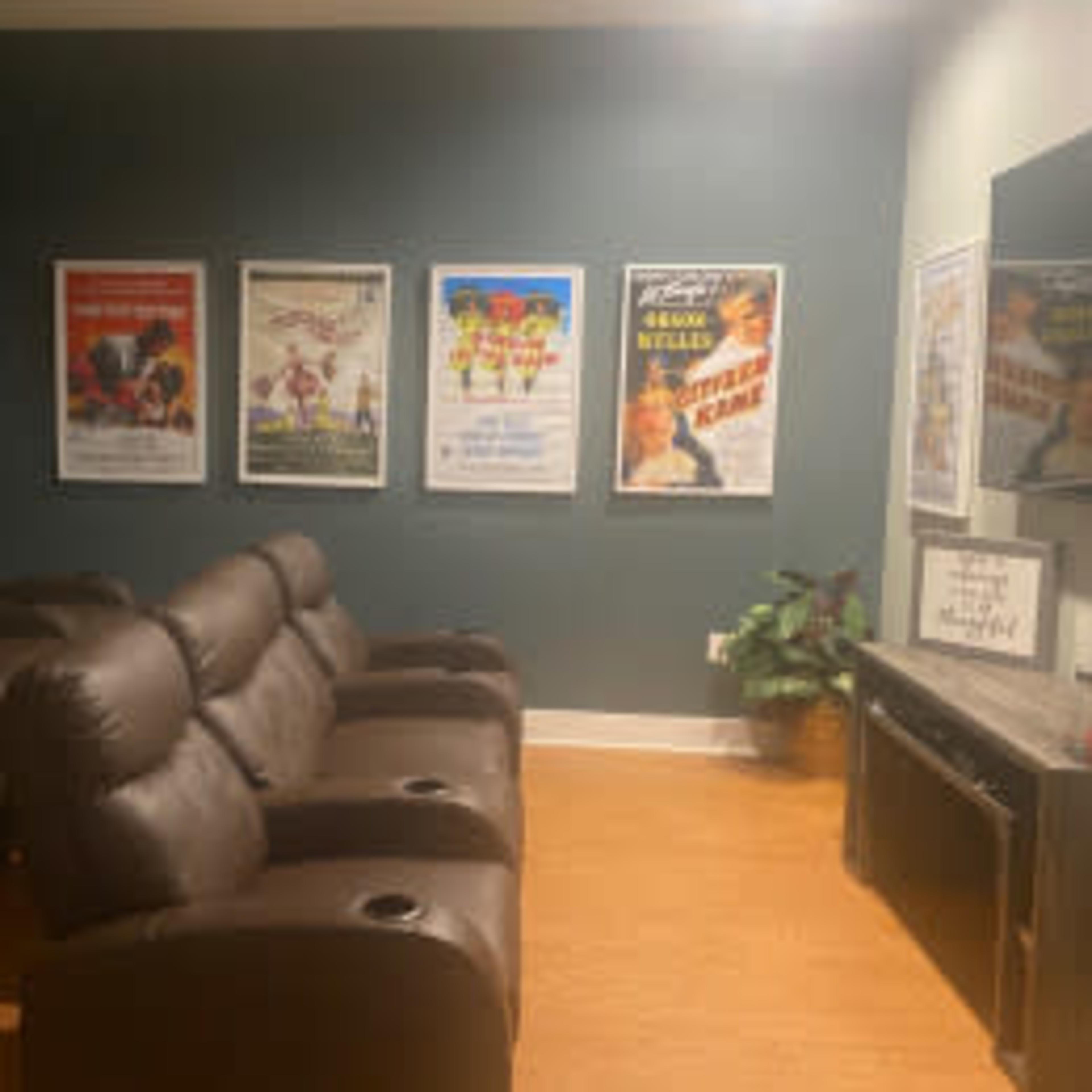 A home theater room with dark recliner seating, movie posters on the walls, and a television mounted on a console.