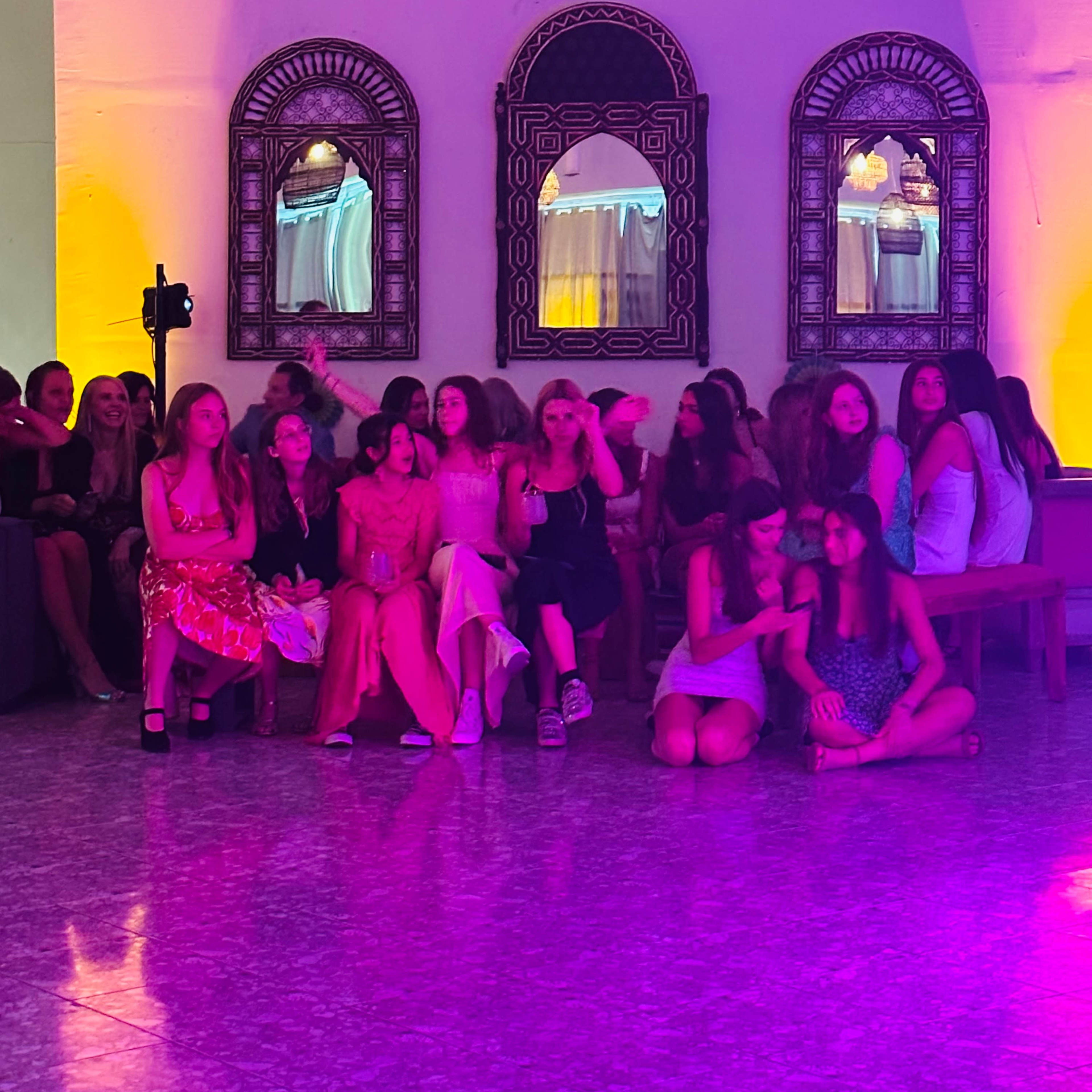 A group of young women sits on benches in a brightly lit room with decorative mirrors on the wall.