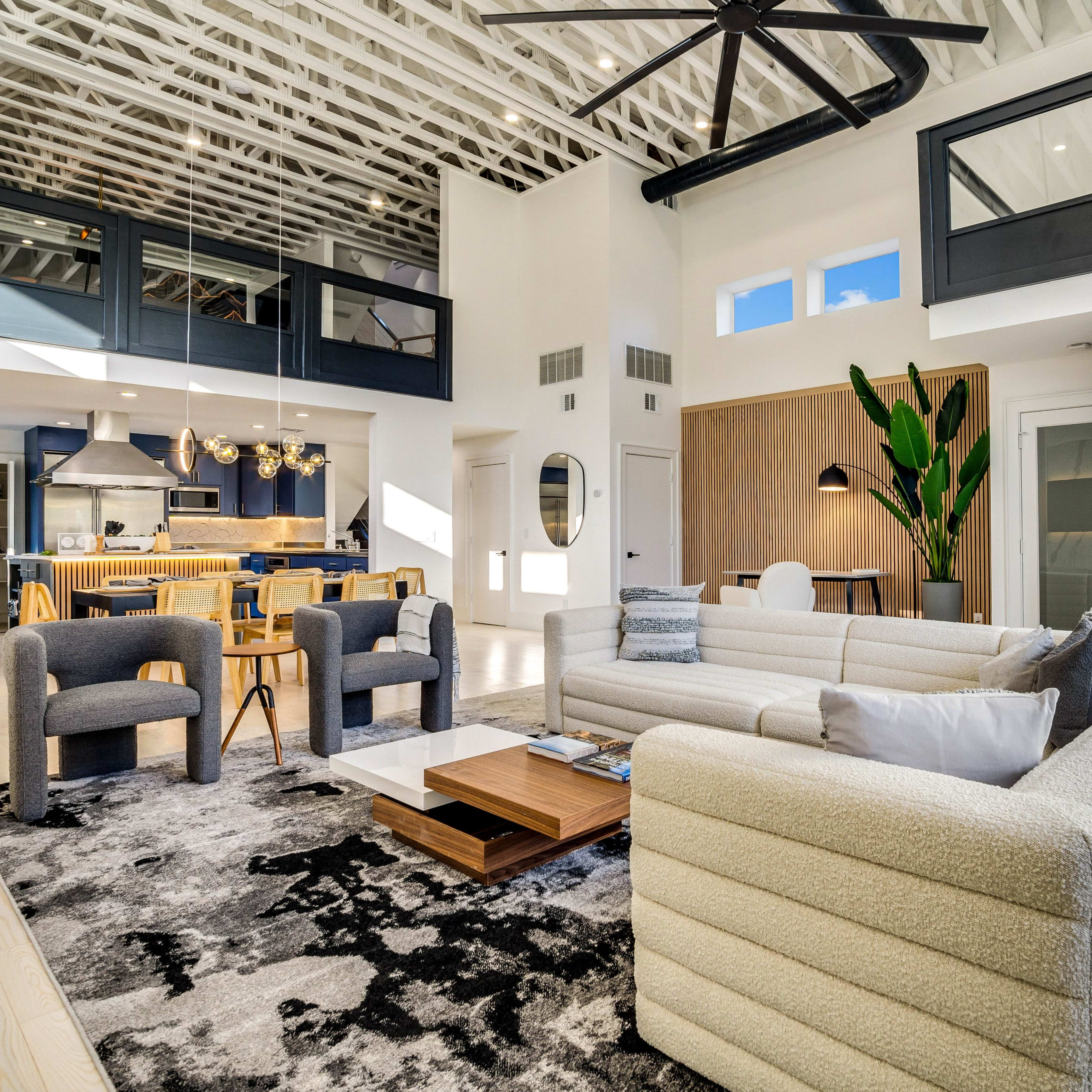 The living room features a spacious layout with a large sectional sofa, two accent chairs, a coffee table, and a tall indoor plant, all set on a patterned rug beneath a high ceiling with exposed beams.