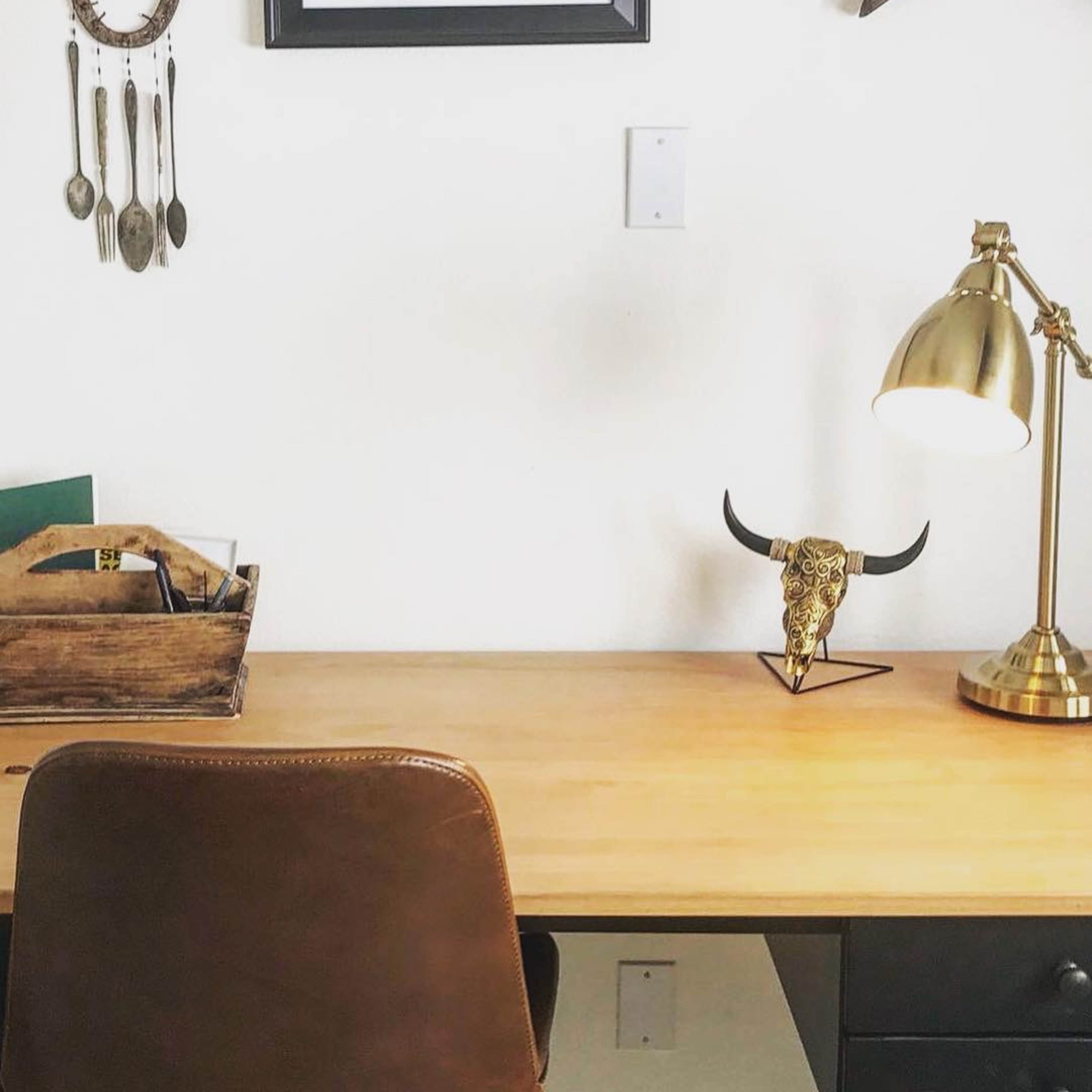 A wooden desk is adorned with a gold lamp and a decorative bull skull, with a framed picture and wall decor in the background.