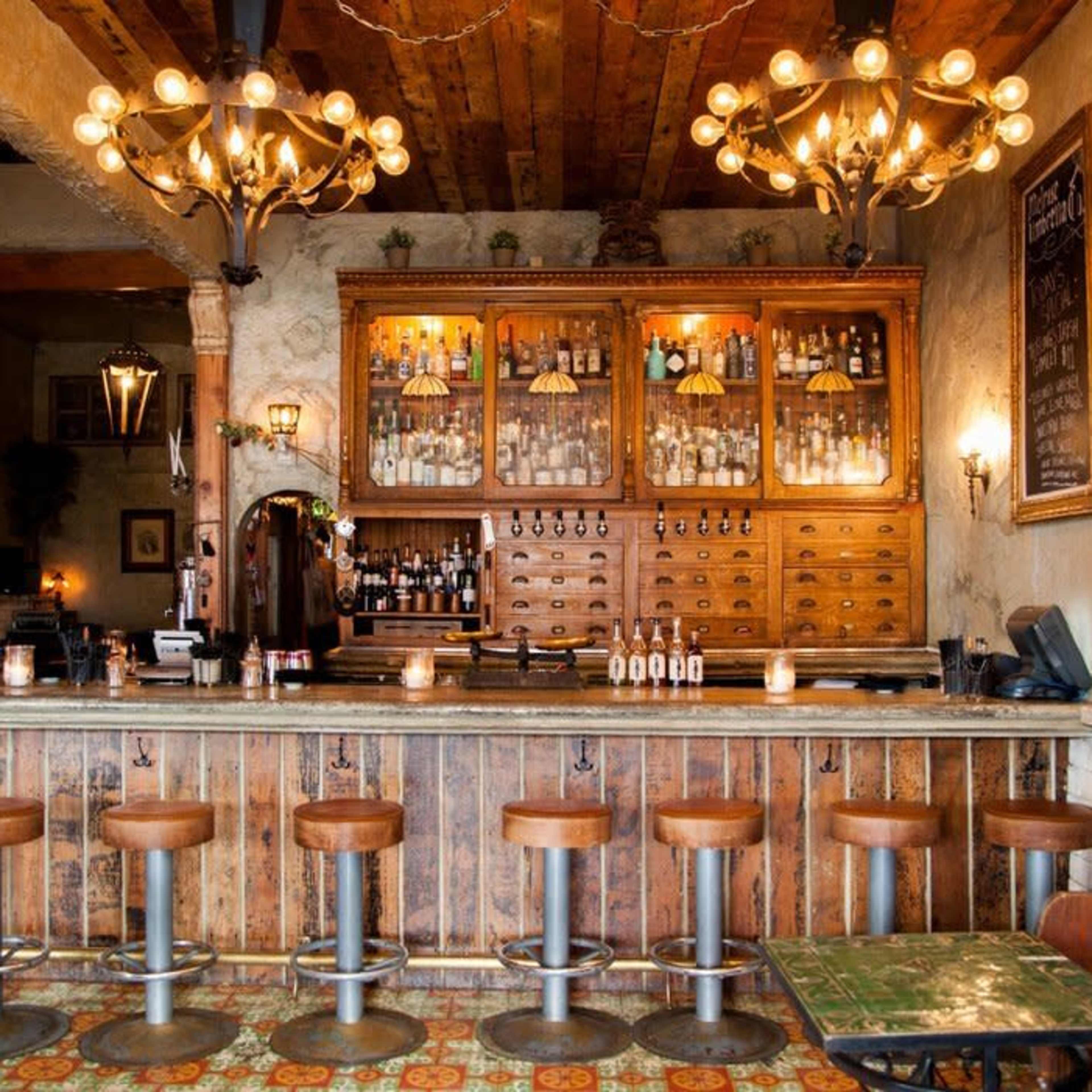 A wooden bar area with high stools and a decorative shelving unit displaying various bottles, illuminated by ornate chandeliers.