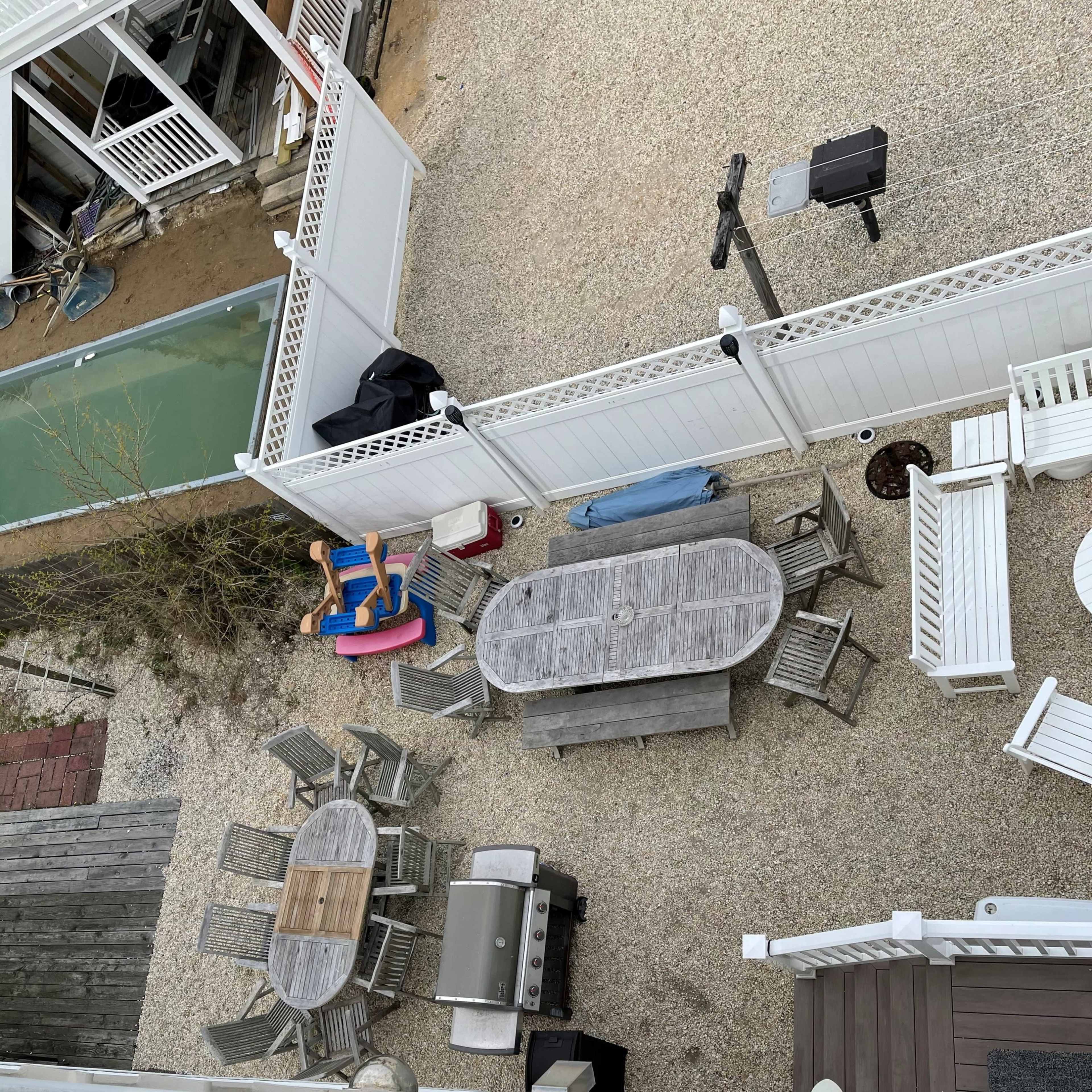 A gravel outdoor area with various furniture, including tables, chairs, and a grill, surrounded by white fences.