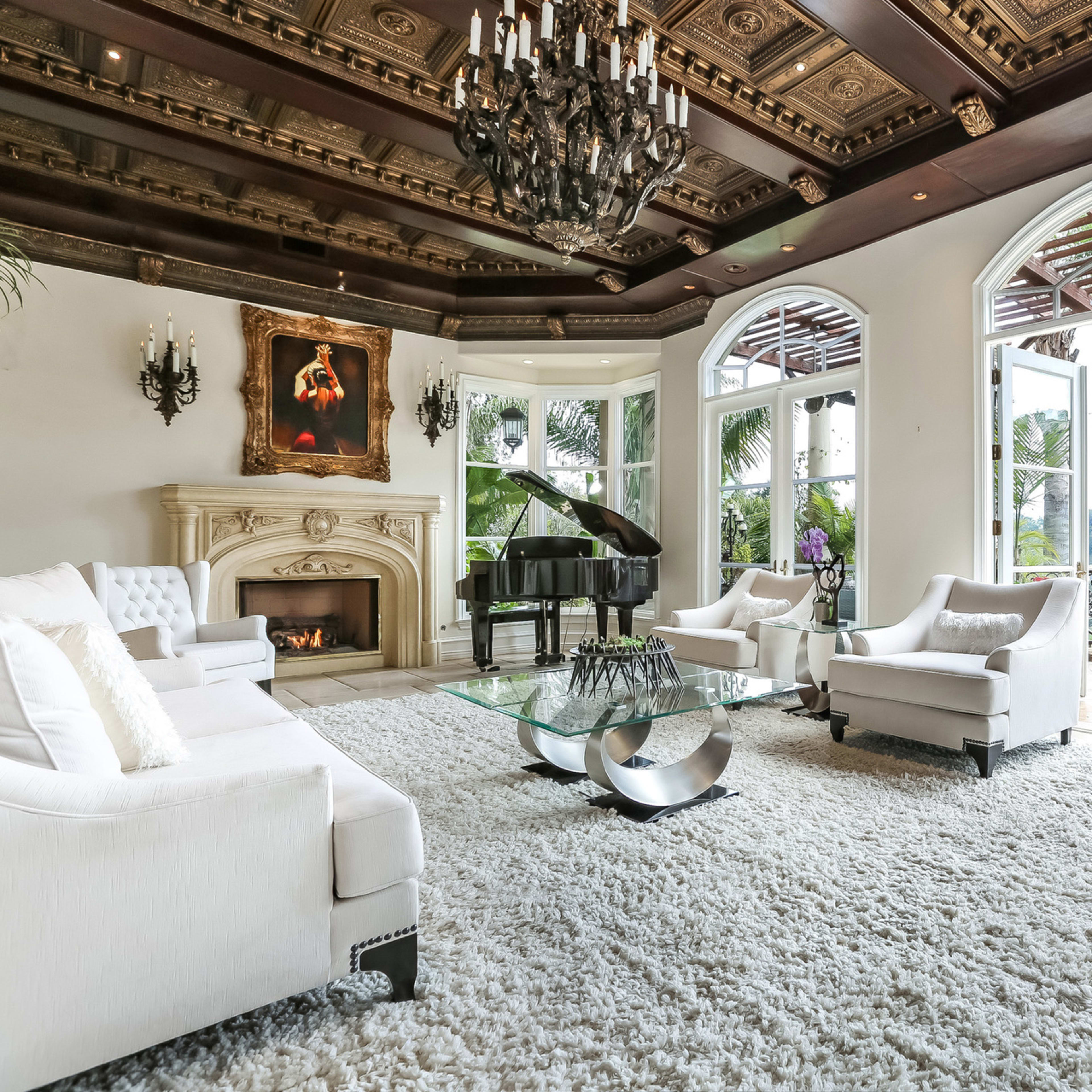 The room features a grand piano beside a fireplace, with large windows offering a view of the outdoor landscape and a plush area rug covering the floor.