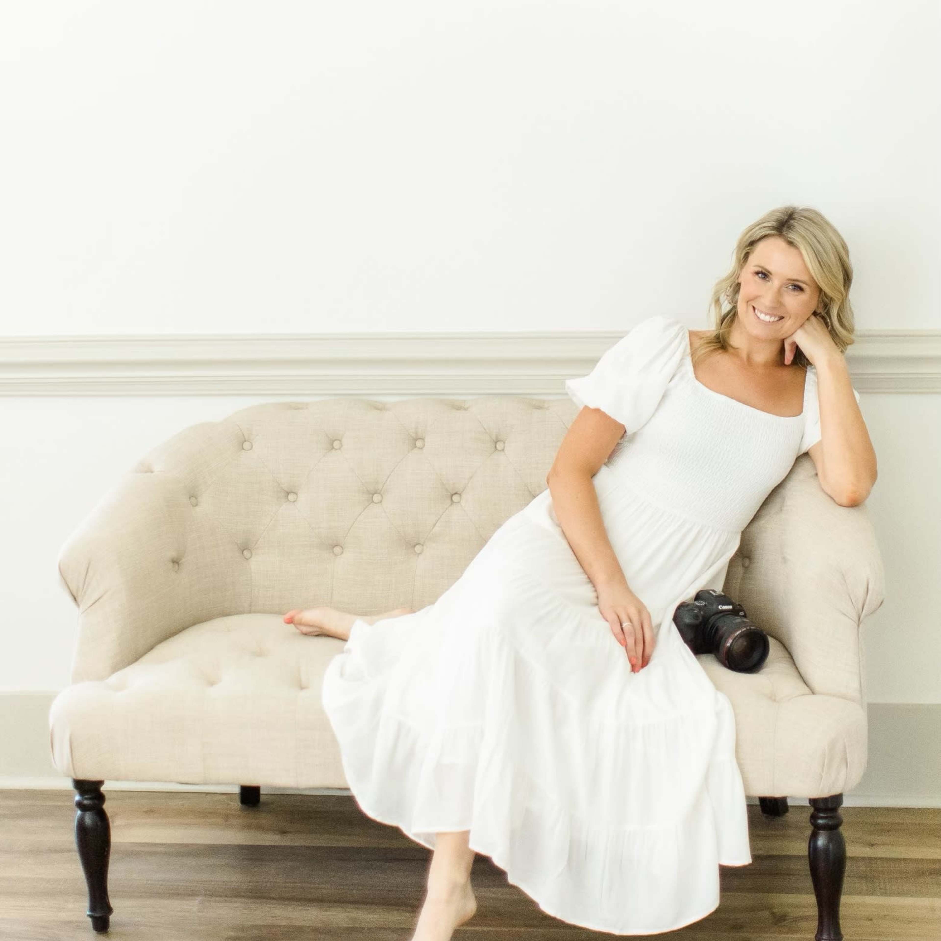 A woman in a white dress sits on a tufted beige couch with a camera resting beside her.