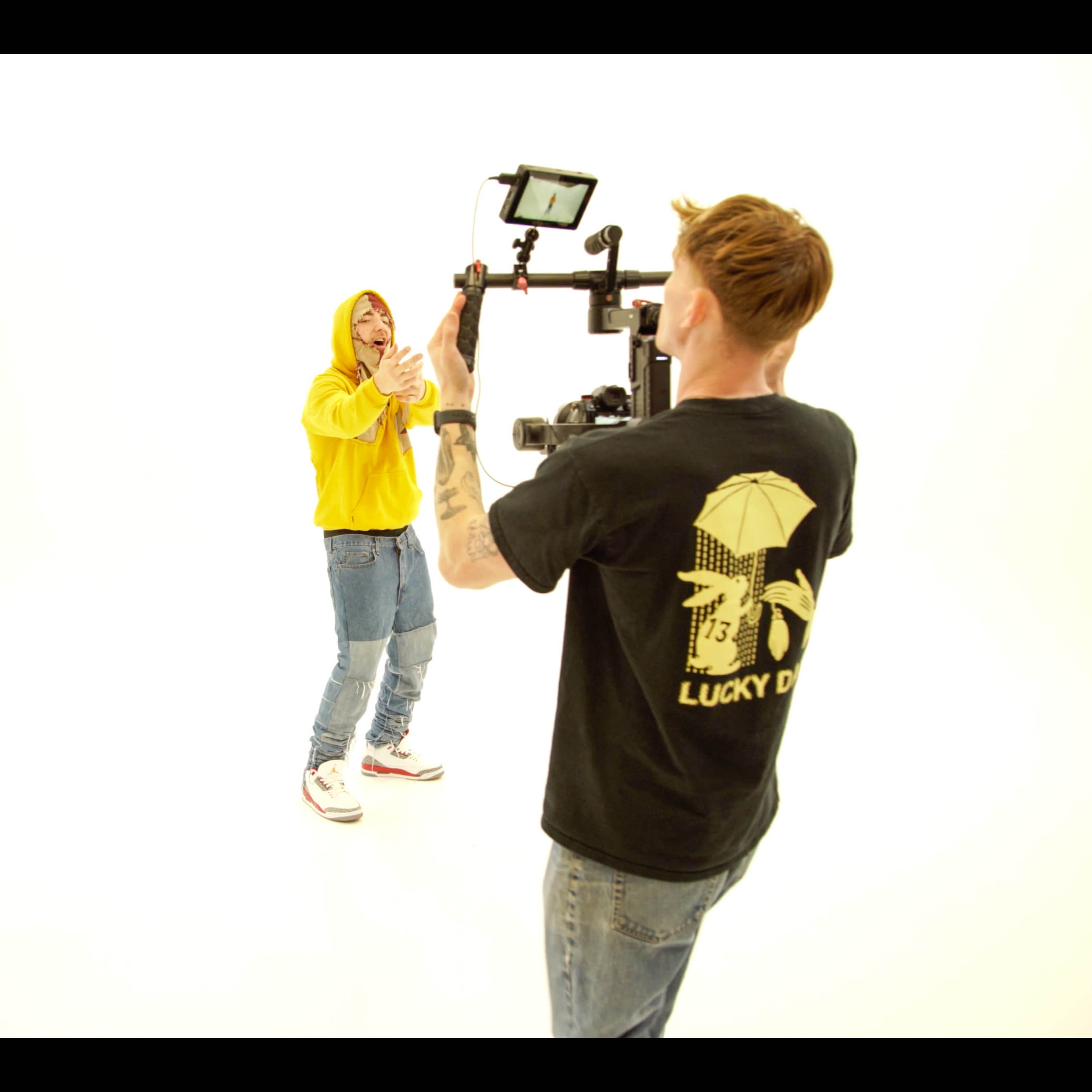 A person in a yellow hoodie performs in front of a camera being operated by another individual wearing a black t-shirt in a white studio setting.