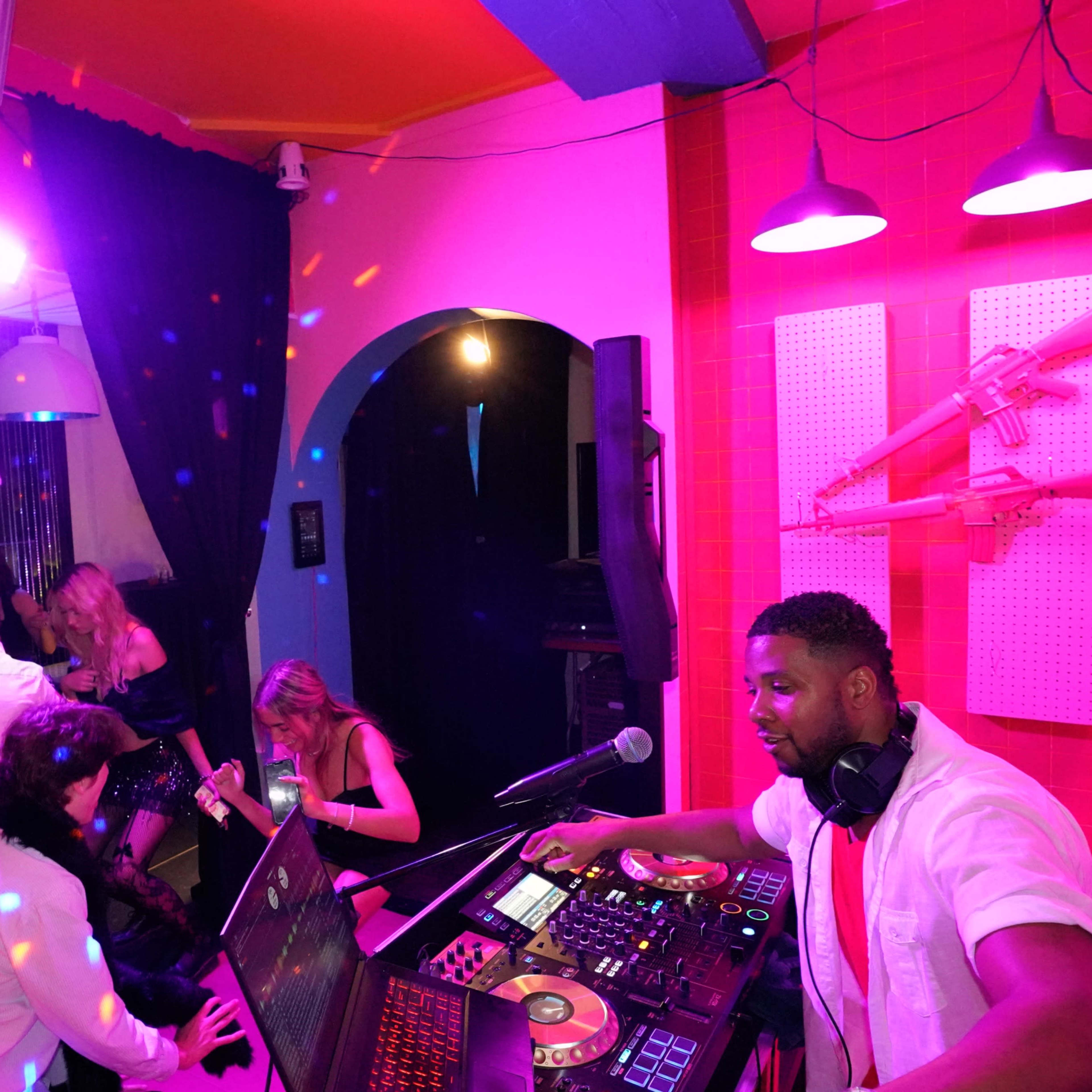 A DJ operates a sound mixer while guests dance and socialize in a vibrant nightclub setting.